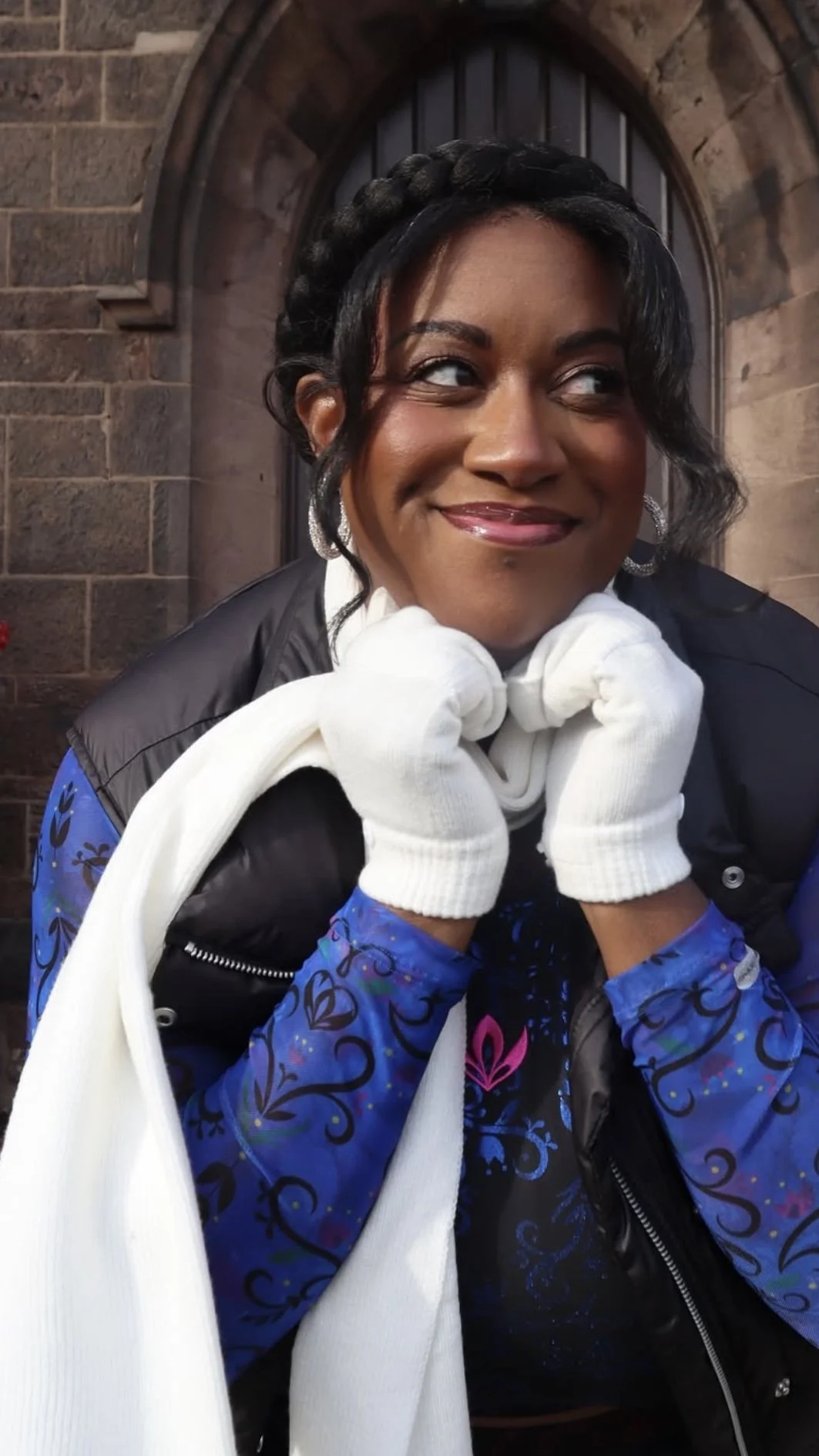 A smiling woman with dark hair styled in curls, wearing earrings, a black vest over a colorful long sleeve shirt, and white gloves, resting her chin on her hands in front of a brick archway.