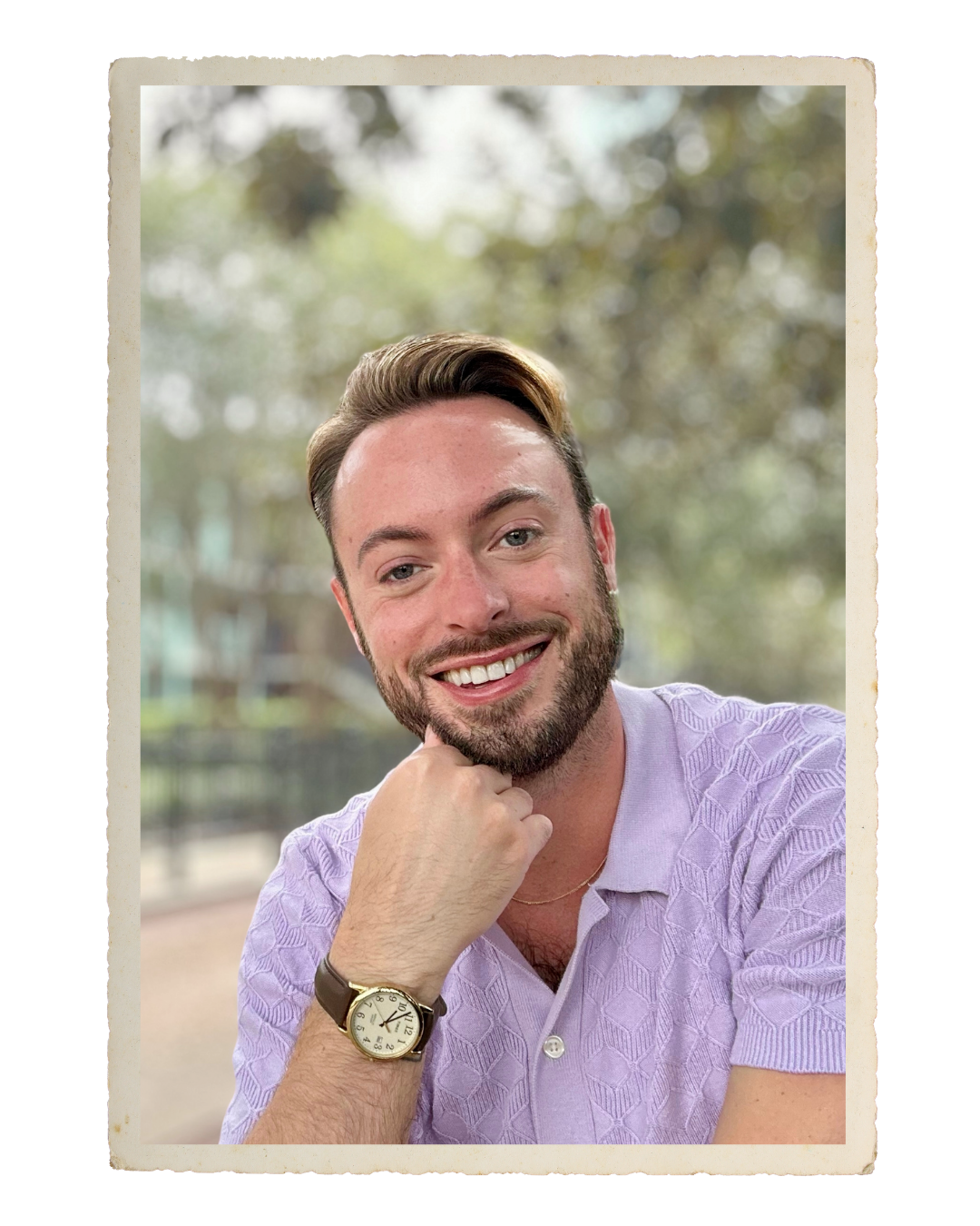 A smiling young man, a personal stylist and wardrobe planner, with a beard and styled hair, wearing a lavender button-up shirt and a watch, sitting outdoors with a blurred background of trees and buildings.