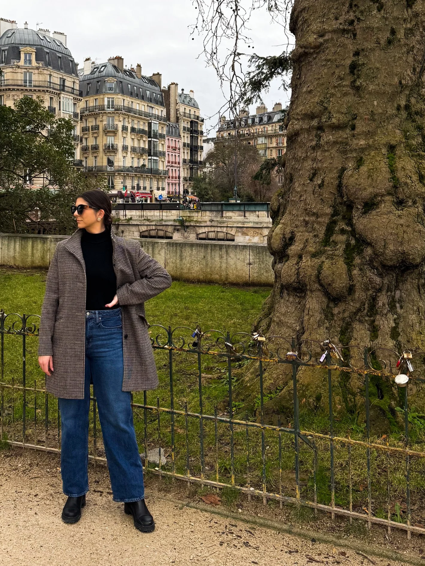 A woman with dark hair, wearing a wardrobe planned outfit including sunglasses, a black turtleneck, and a checkered coat standing with her hand on her hip near a large tree in a Parisian park.