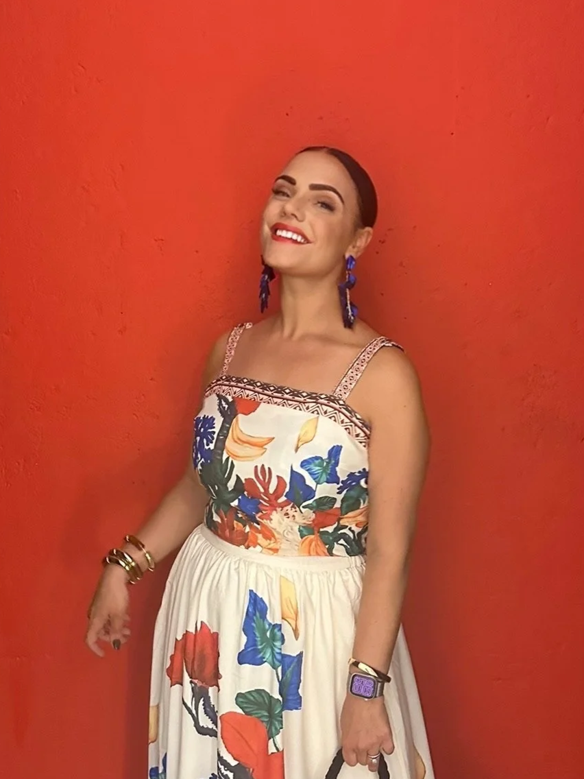 A woman with dark hair styled in a bun, wearing a colorful vacation outfit, a floral dress and large blue earrings, standing against an orange wall, smiling and winking at the camera.
