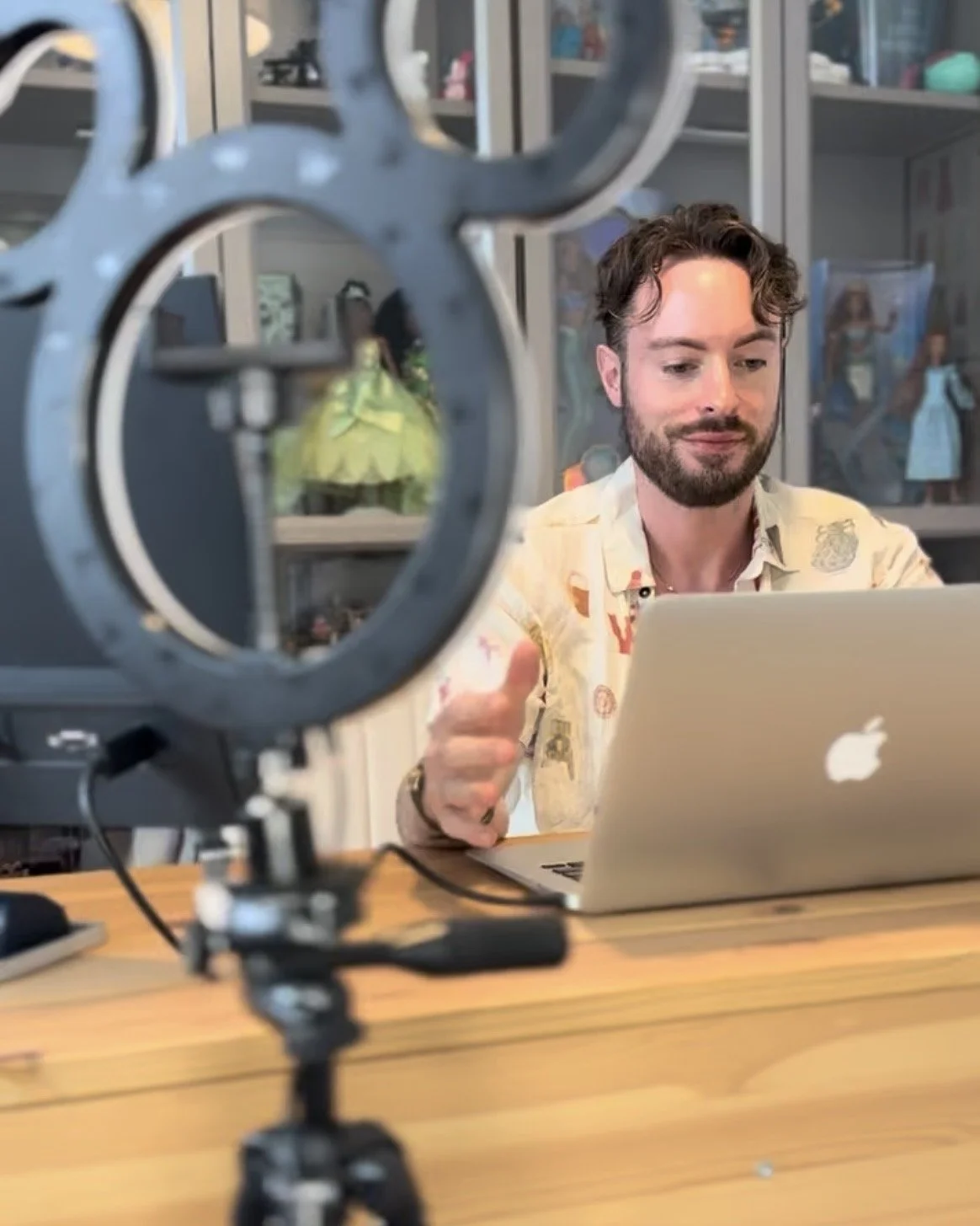 A personal stylist, a man, working on a laptop behind a ring light, with shelves filled with collectibles in the background.