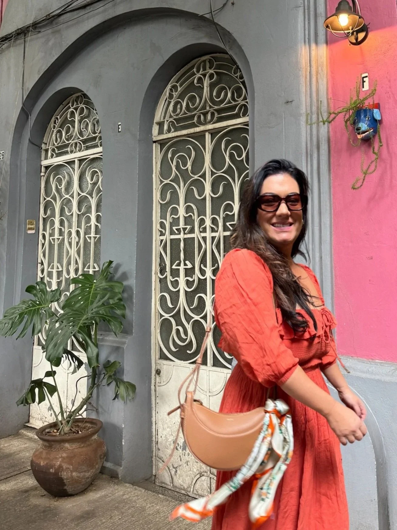 A woman in a vacation outfit: an orange dress with sunglasses and a beige handbag standing in front of a gray building with ornate metal gates.