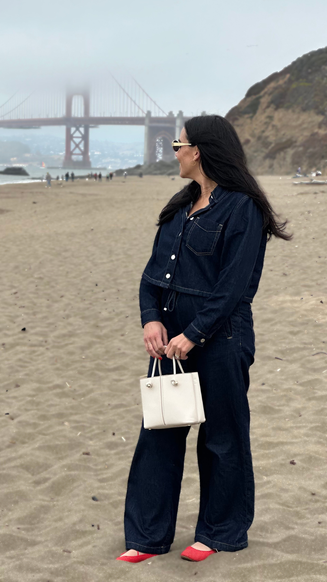 Woman standing on a sandy beach near San Francisco, wearing a personally styled outfit: dark jeans and a denim jacket, carrying a white handbag, with the Golden Gate Bridge visible in the background.