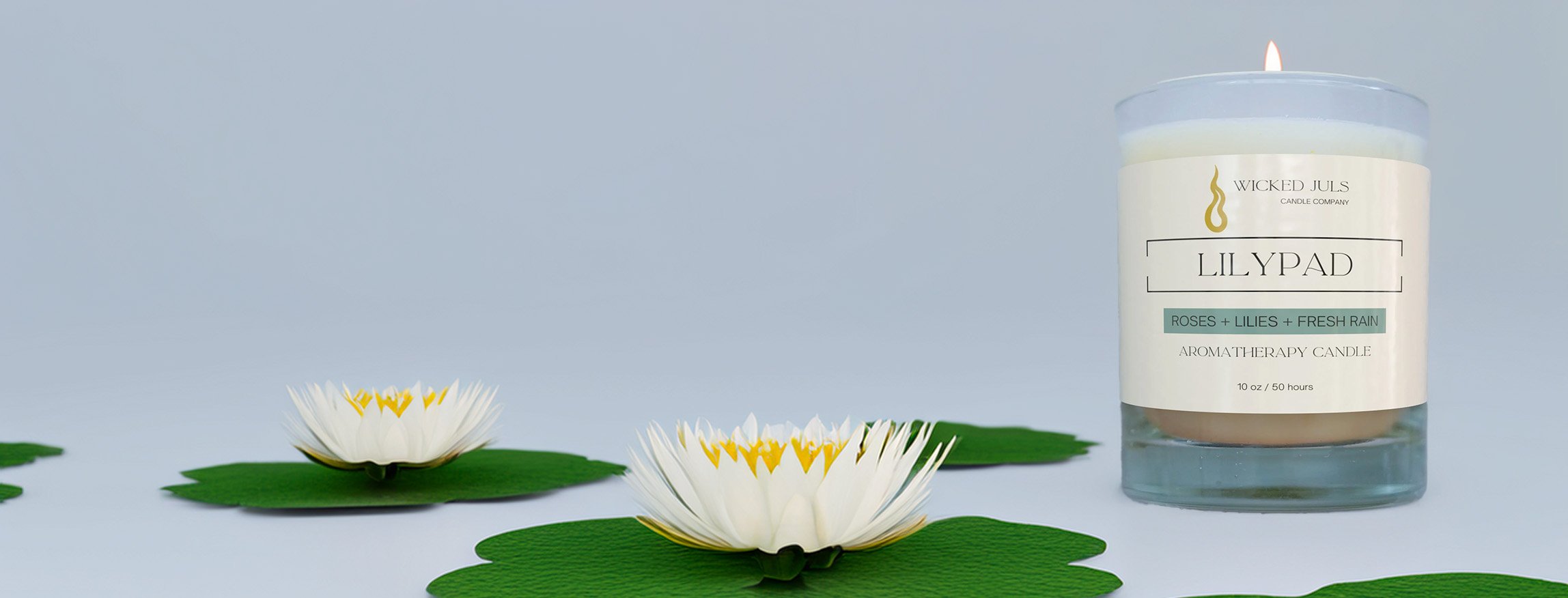 A lit aromatherapy candle labeled 'Lillypad' with roses, lilies, and fresh rain scent, surrounded by water lilies on a white background.