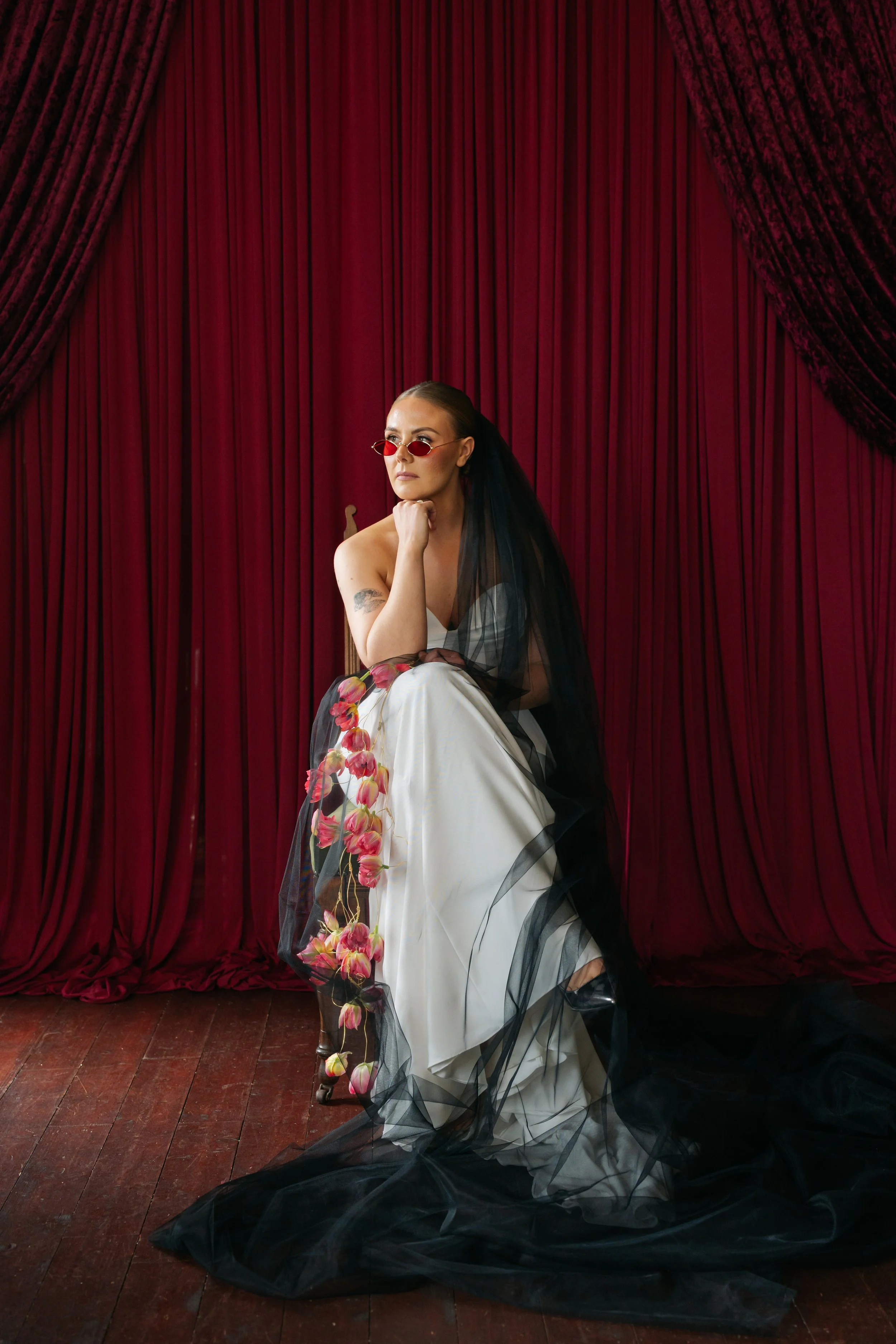 Bride sitting in front of red velvet stage curtains, wearing a white dress with black sheer veli, red sunglasses, and drape floral bouquet. Drapes styled by alternative wedding stylist Cherry on Pop in Lanarkshire