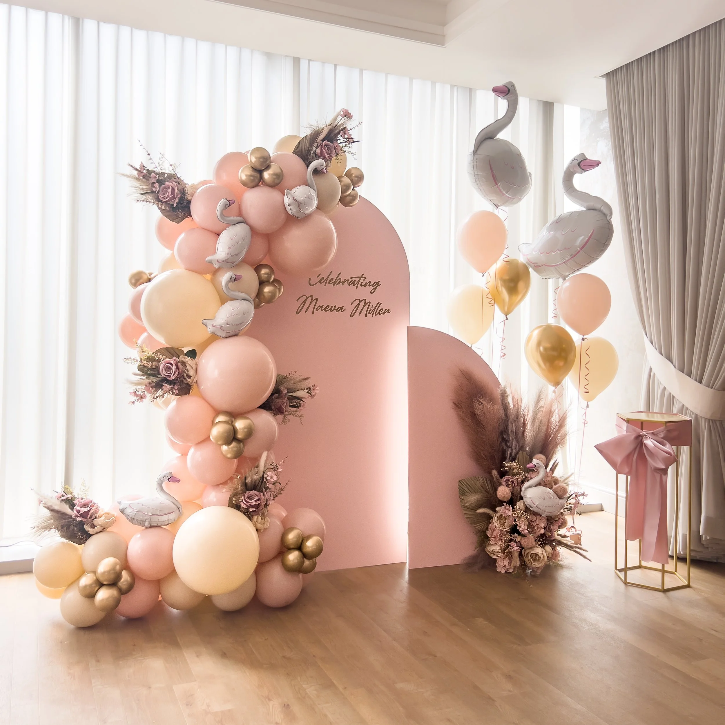 Decorative setup with balloons and flowers celebrating Maeva Mullen with pink, cream, and gold balloons, swan-shaped balloons, a pink backdrop, and floral arrangements.