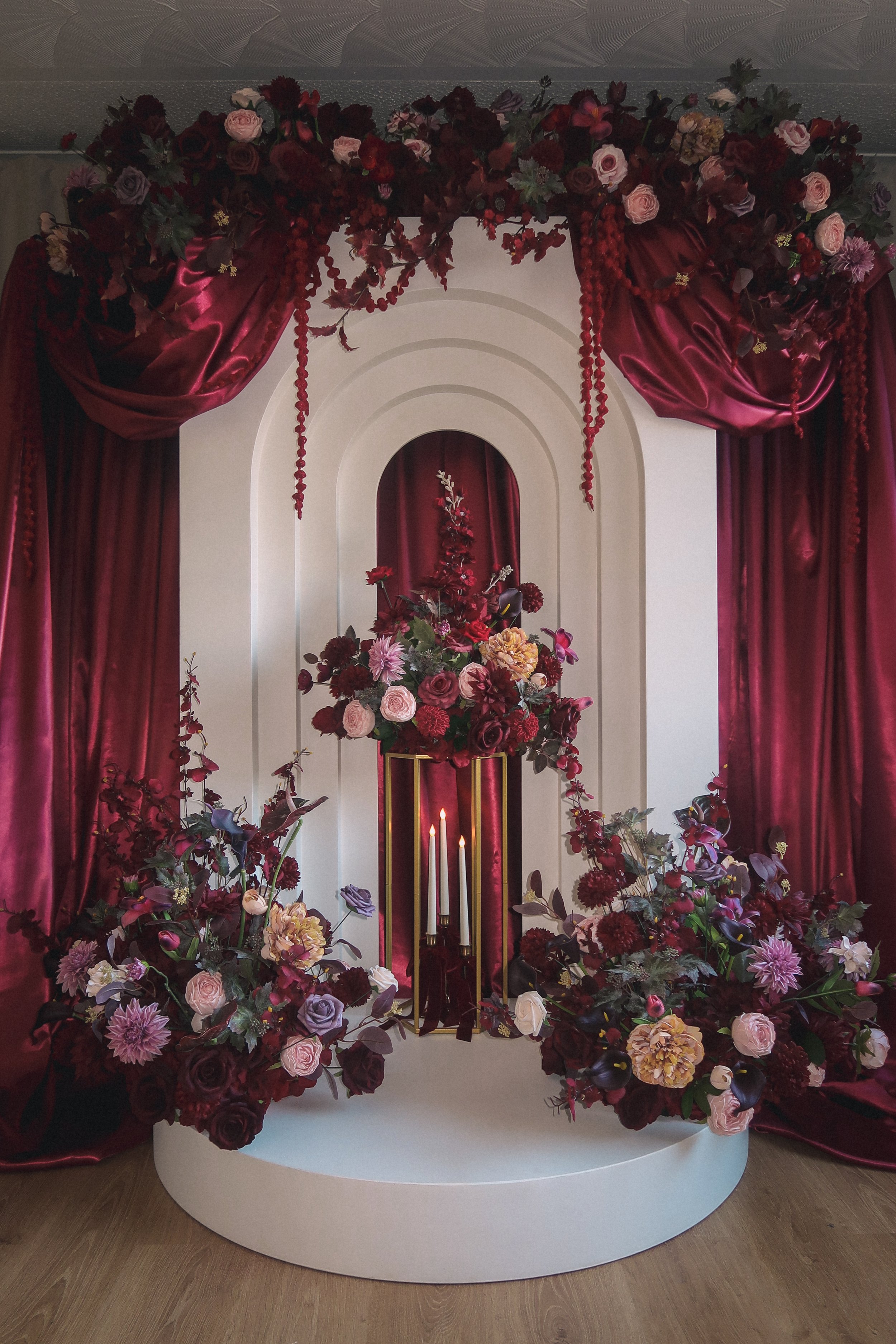 An elaborate floral arrangement with deep red, pink, and purple flowers, set against white and burgundy draped curtains and a white backdrop with arches, featuring candles and gold accents.