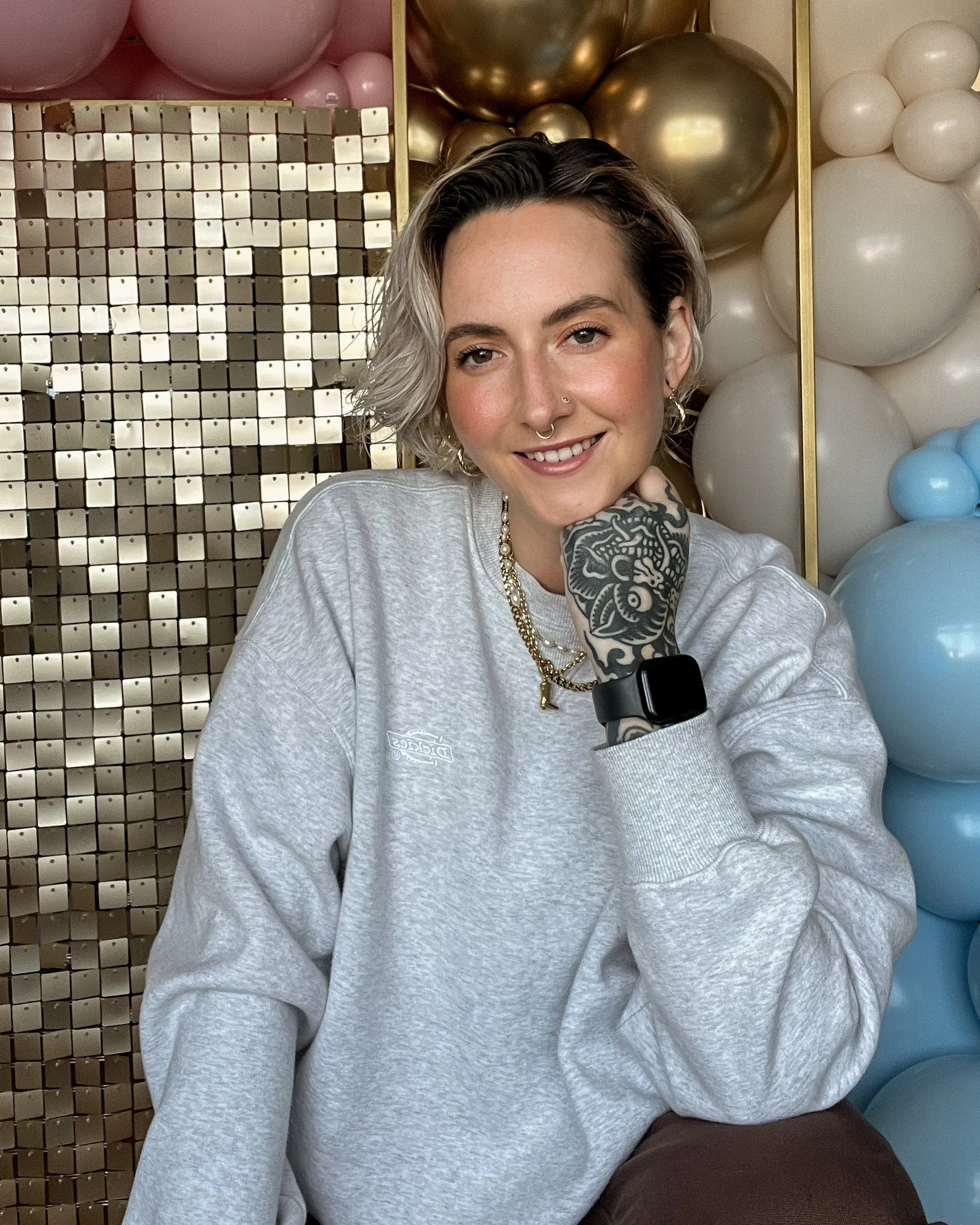 Young woman with short, styled, gray and black hair, wearing a gray sweatshirt, layered necklaces, and a smartwatch, sitting in front of balloons and metallic decorations and smiling at the camera.