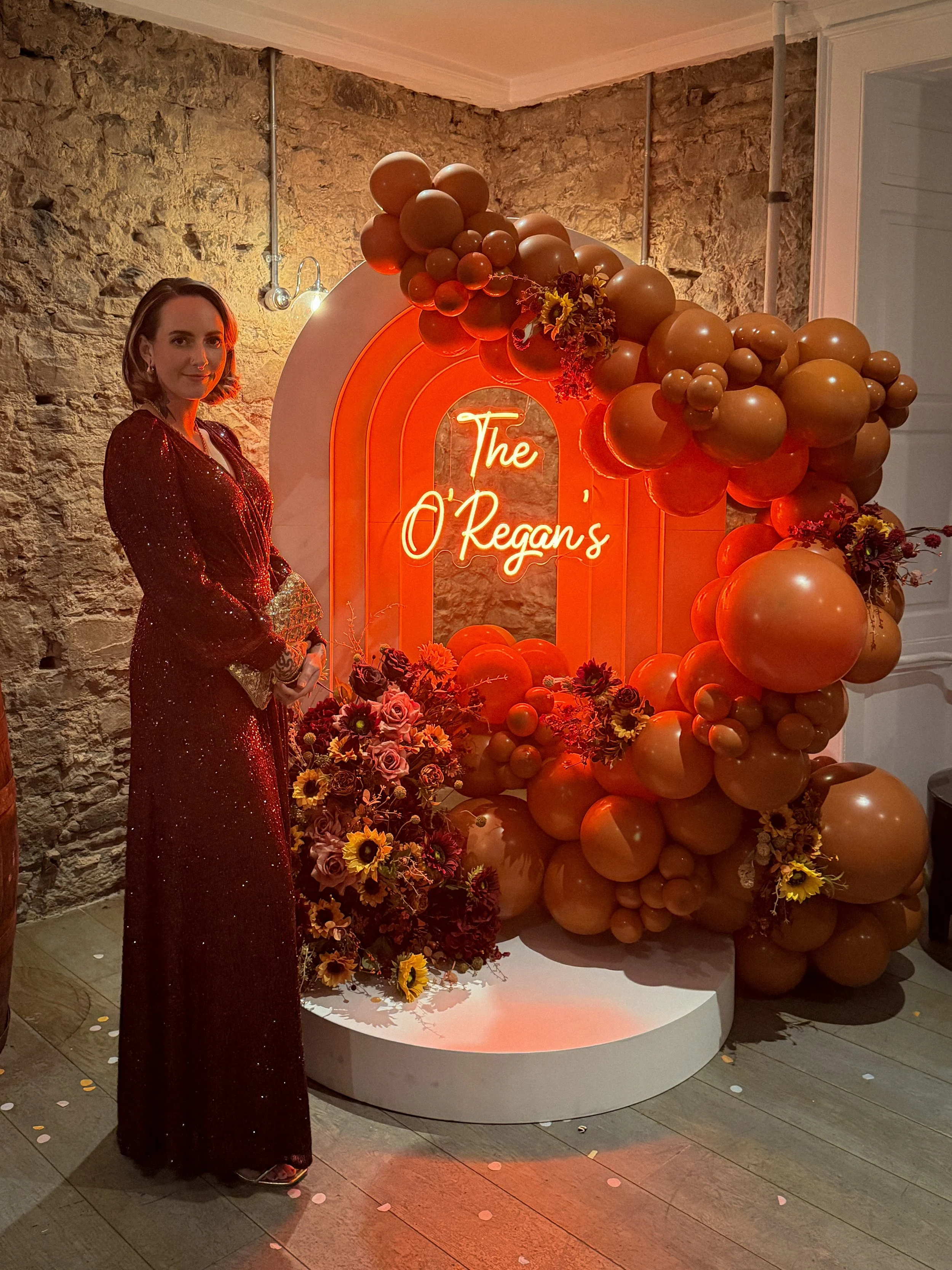 Woman in a red shimmering dress standing next to a decorative backdrop with balloons and floral arrangements that says "The O'Regan's", at an indoor event or celebration.