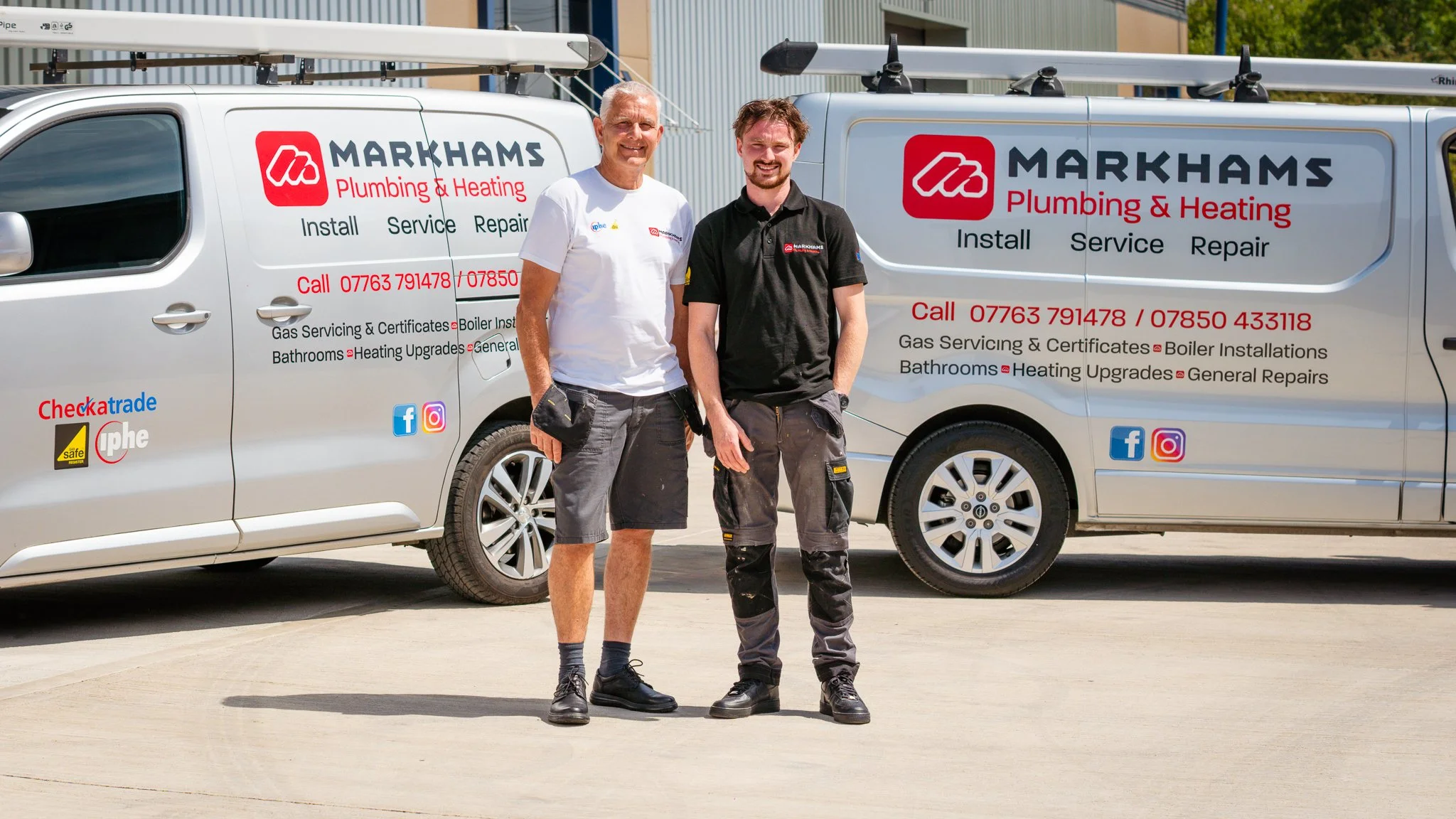 Two men standing in front of branded service vans for Markhams Plumbing & Heating. One is wearing a white t-shirt and shorts, the other a black polo and work pants. Both are smiling.