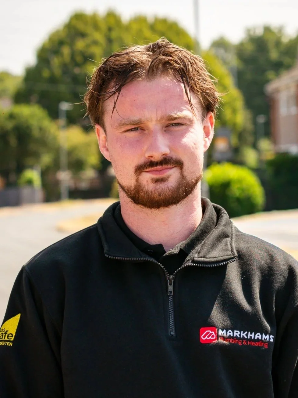 A man with light skin, brown hair, and a beard smiling outdoors with greenery in the background. He is wearing a black collared shirt with a logo on the chest that reads "Markhams Plumbing & Heating."
