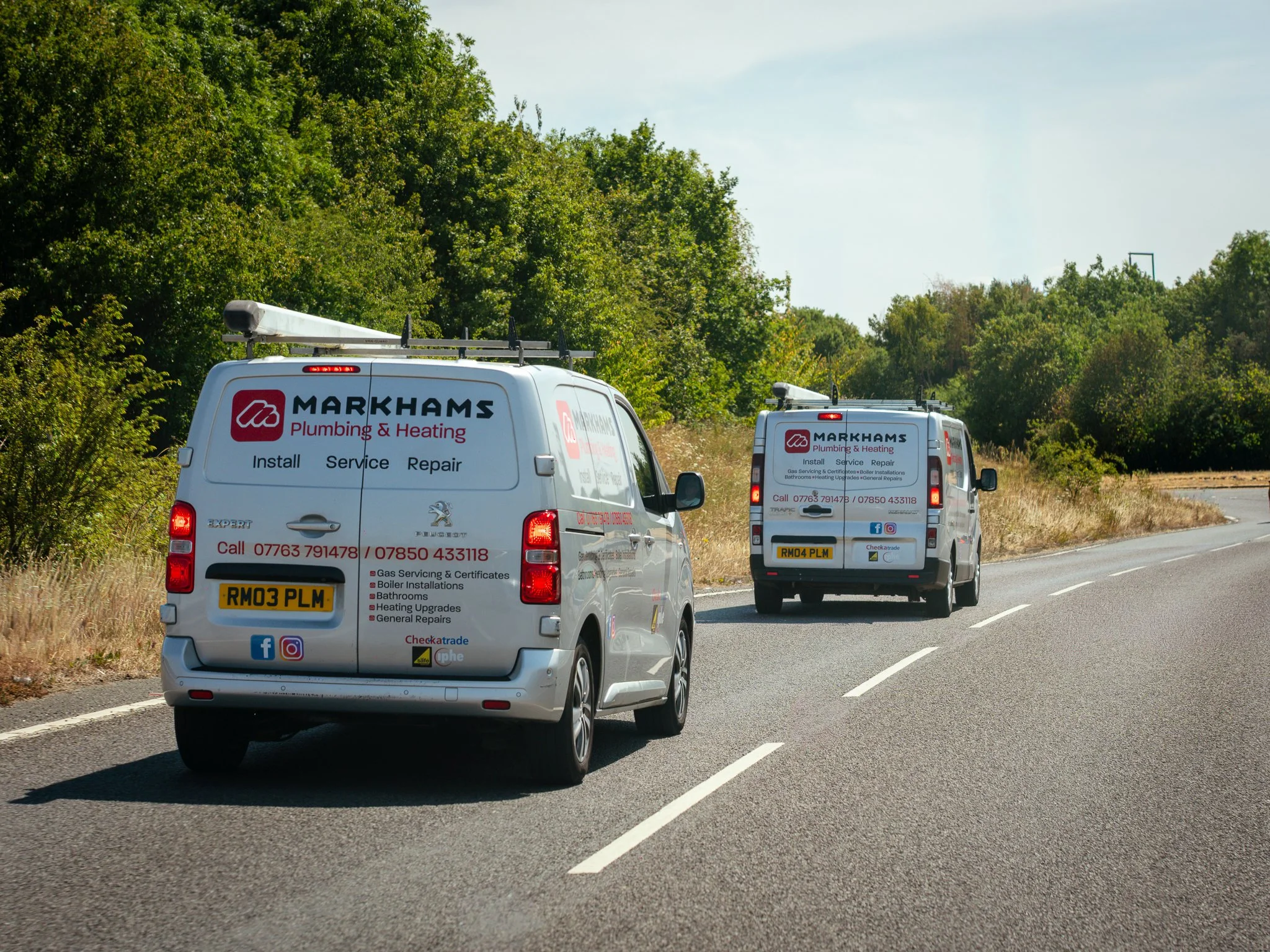 Two white vans with 'Markhams Plumbing & Heating' branding parked on the side of a two-lane road, surrounded by green trees and brush under a blue sky.