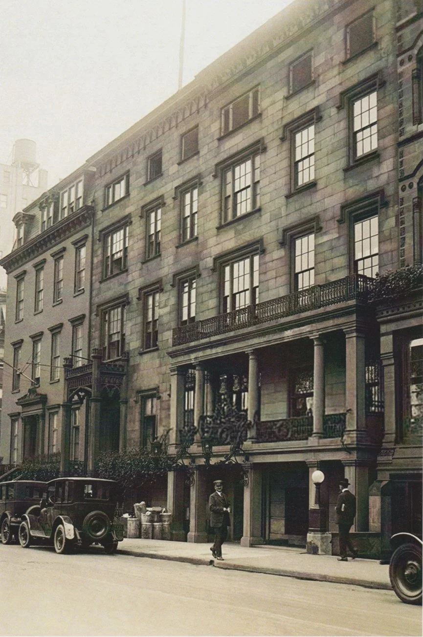 A black-and-white photo of a city street with a multi-story building featuring classic architecture, including columns and detailed window frames. Two men are walking on the sidewalk, and several vintage cars are parked along the street.