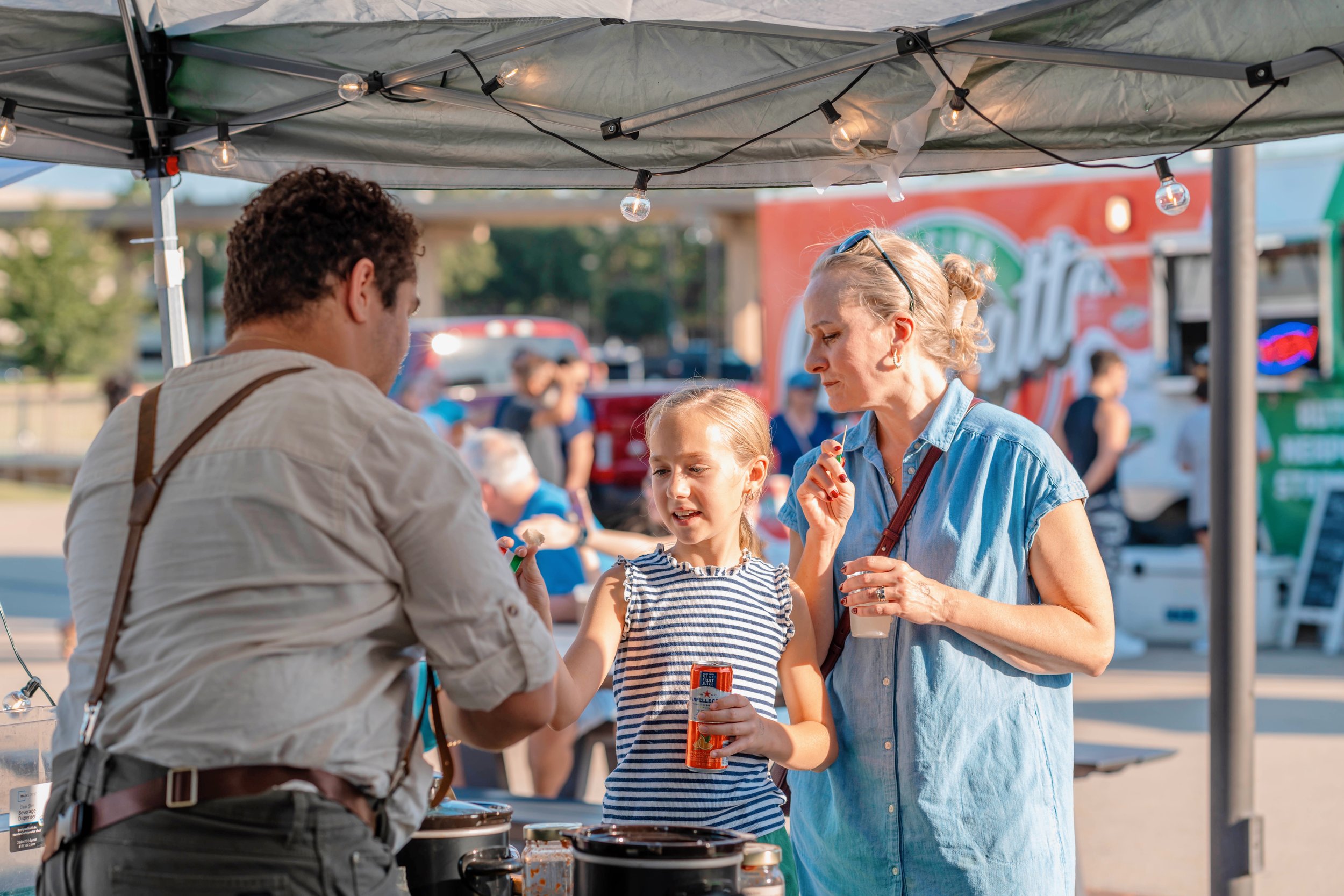 A man serves food samples to a woman and a young girl at an outdoor market under a canopy. The woman holds a small cup and a sampling skewer, while the girl holds a can and engages with the vendor. The setting appears to be a fair or festival, with f