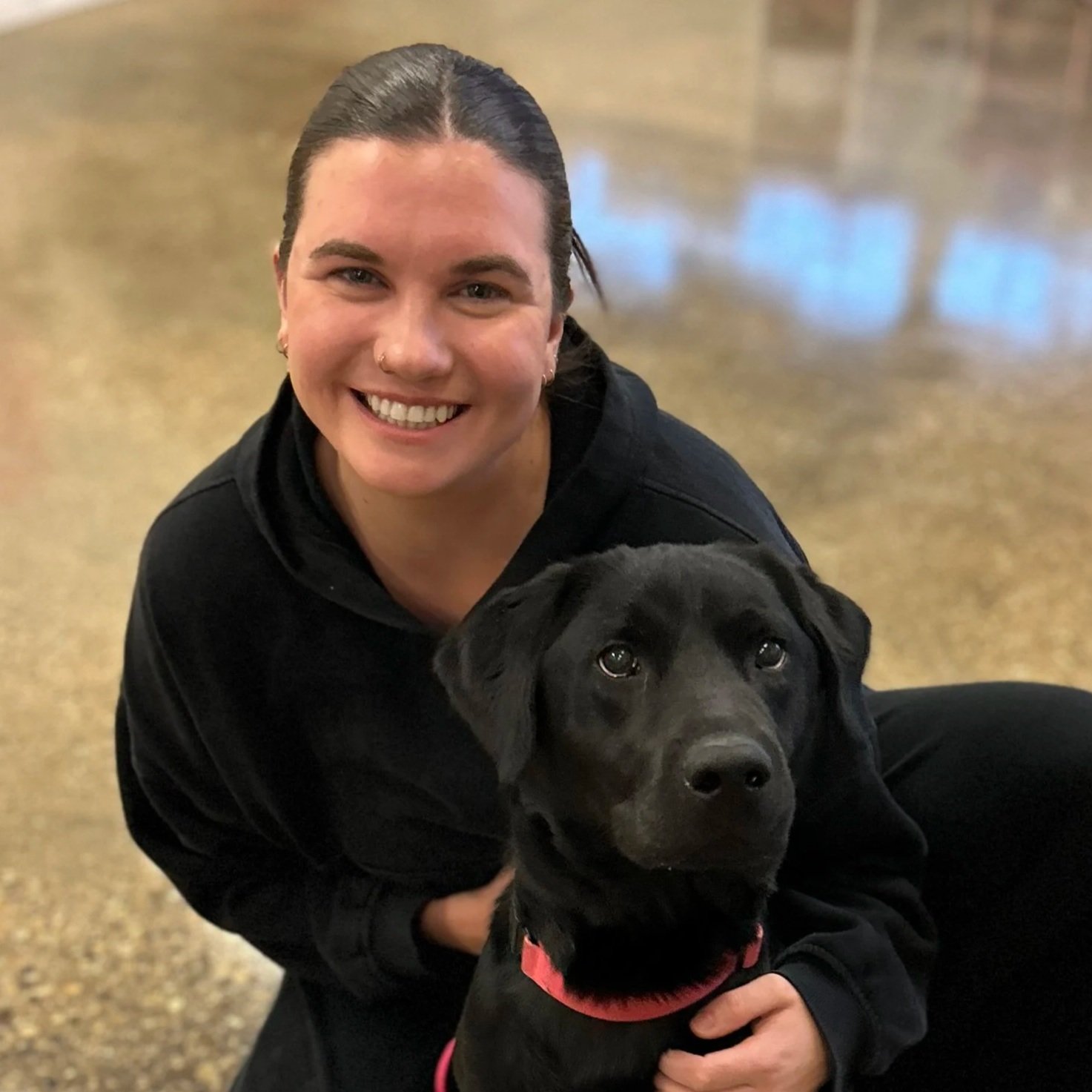 dark haired woman in a black sweatshirt poses with a black labrador retriever