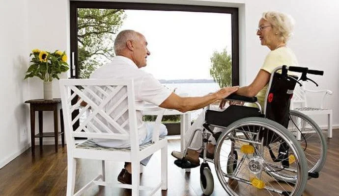 An elderly man and woman in a wheelchair holding hands inside a room with a view of a lake outside the window.
