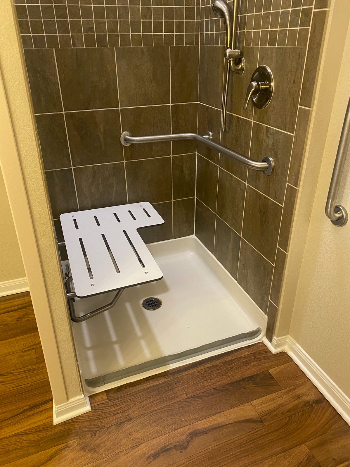 Shower stall with brown tiled walls, grab bars, a shower head, and a foldable shower seat on a white shower base with a drain.