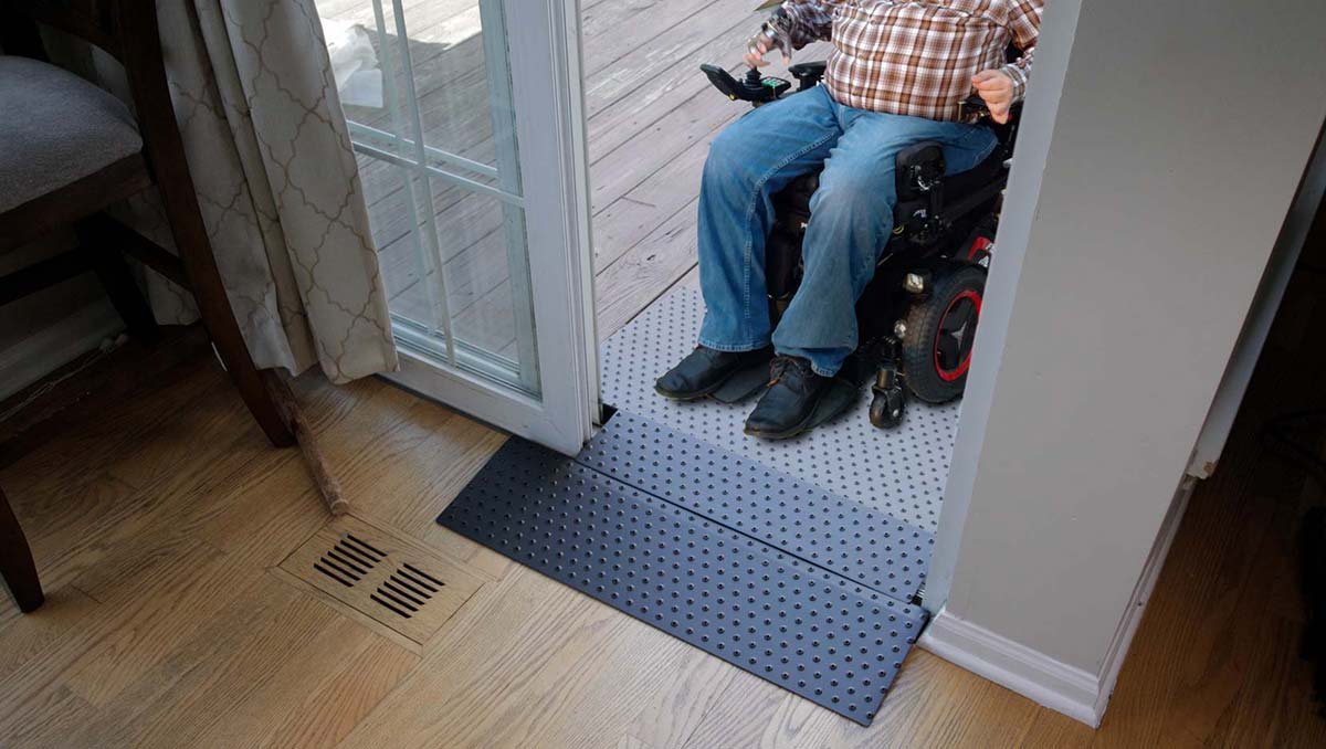 A person in a motorized wheelchair on a deck outside, viewed through a sliding glass door, with a wooden floor and a vent inside.