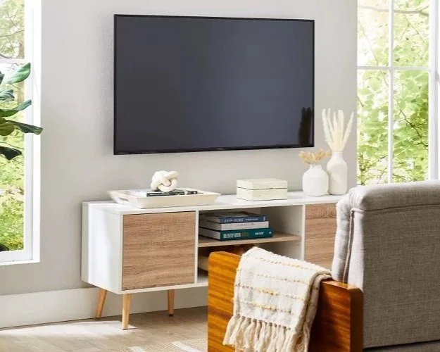 Modern living room with wall-mounted TV, white and wood console table, decorative vases, and armchair with throw blanket, next to windows with greenery outside.