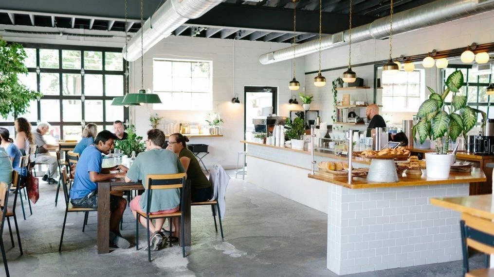 Interior of a bright, modern cafe with customers sitting at tables, a counter with baked goods, and large windows providing natural light, decorated with plants and pendant lights.