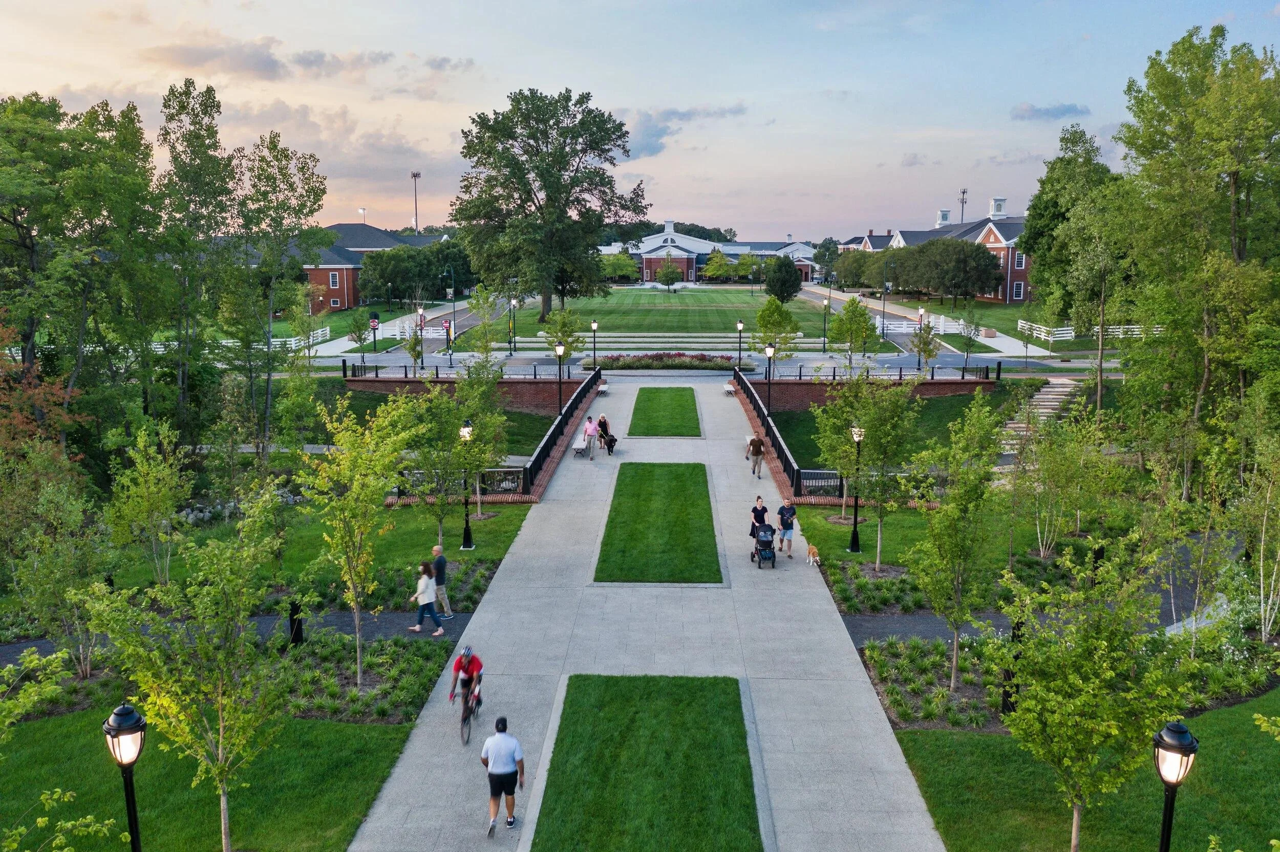 A scenic park with a wide walkway lined with lampposts, trees, and benches, leading to a large building in the distance with a grassy area and a parking lot, with several people walking, biking, and pushing strollers.