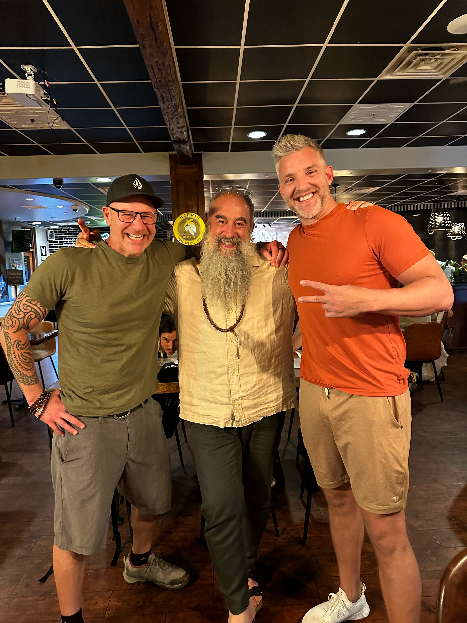 Three men are smiling and posing for a photo in a restaurant. The man on the left has blonde hair, is wearing an orange t-shirt and shorts and making a peace sign. The man in the center has long gray hair, a beard, is wearing a beige linen shirt and 