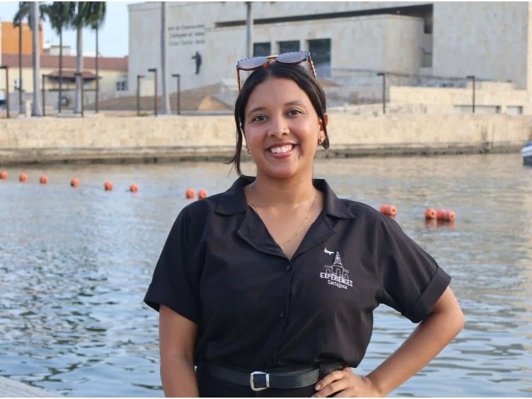 Young woman smiling outdoors near a body of water, wearing a black shirt with a logo, sunglasses on her head, and her hand on her hip, with buildings and trees in the background.