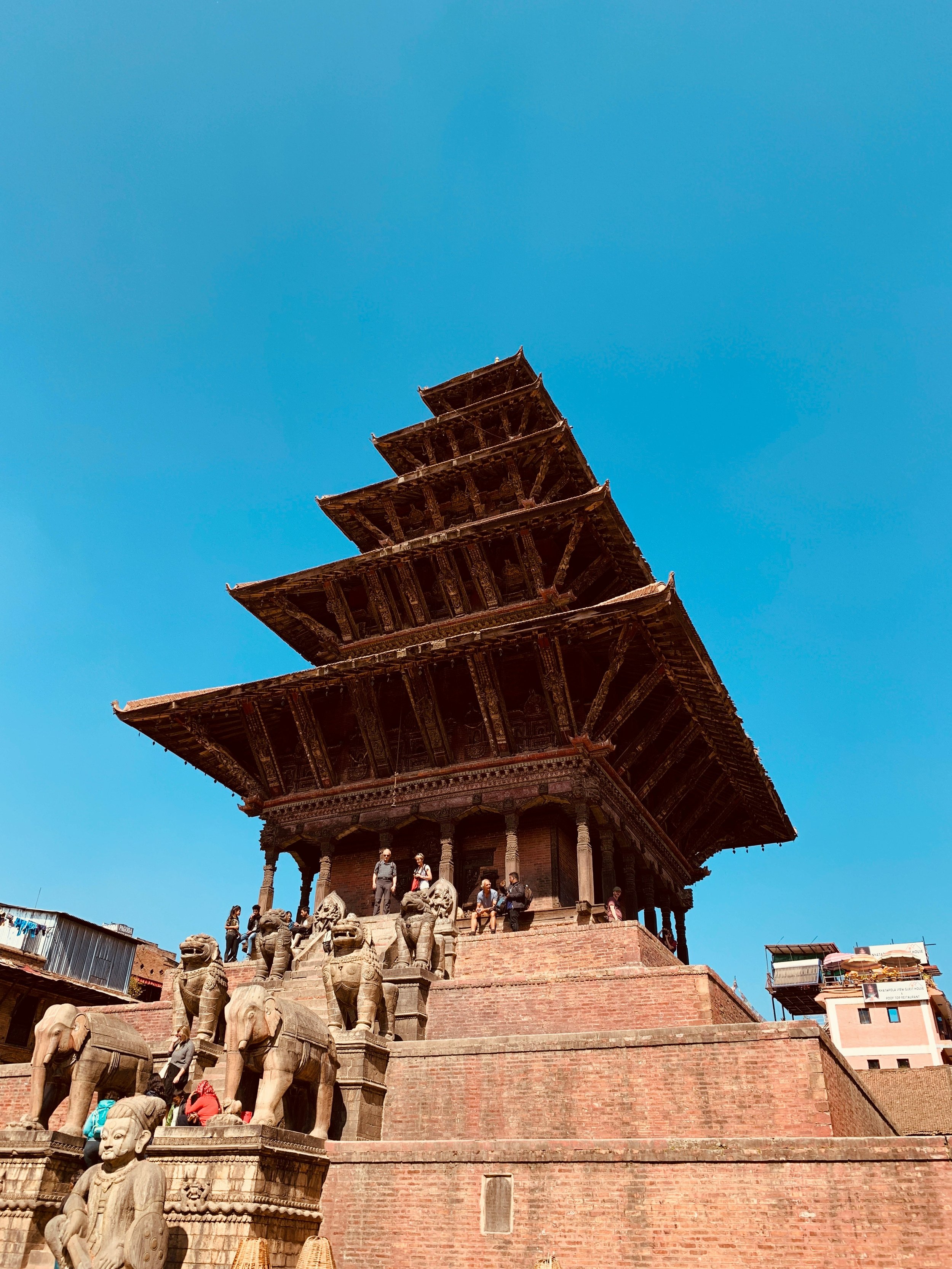 Ancient tiered temple with wooden roof, stone stairs, elephant statues, and visitors, under a clear blue sky.