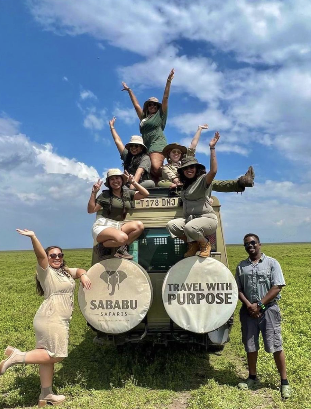 A group of tourists enjoying a safari adventure, sitting and posing on and around an old vehicle in a grassy plain under a bright blue sky with scattered clouds. They are wearing safari hats and casual outdoor clothing, smiling and making playful poses.