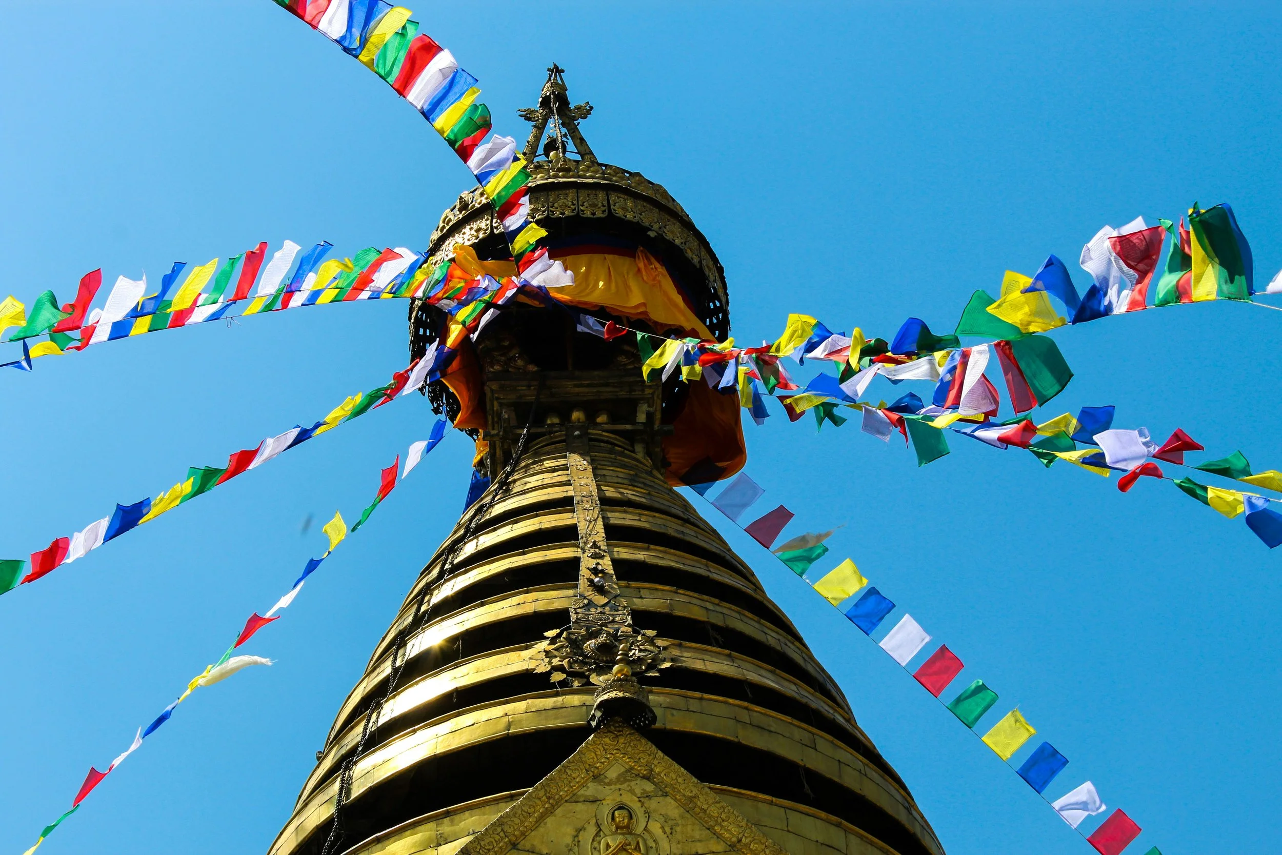A close-up view of a golden stupa with colorful prayer flags attached, set against a clear blue sky.