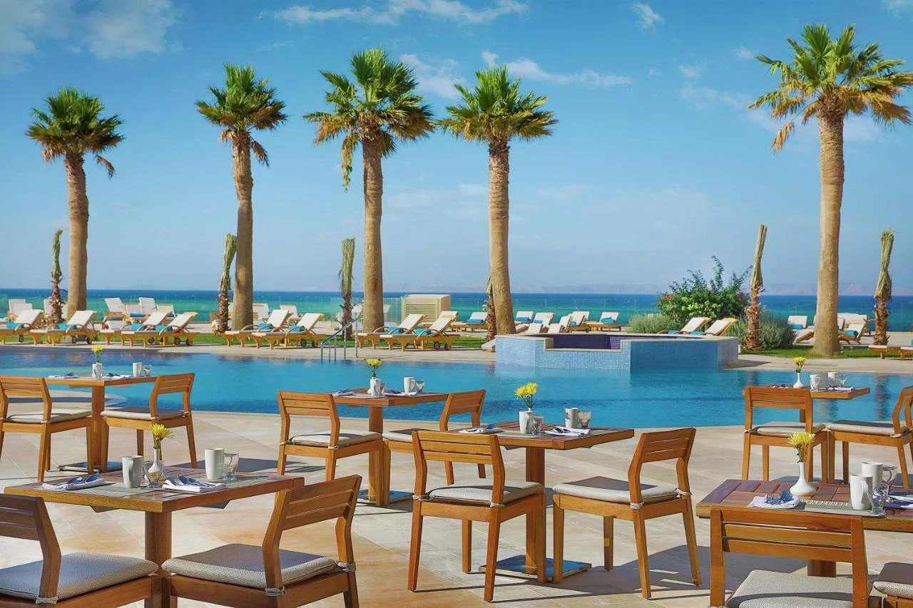 Poolside dining area with wooden tables and chairs, set with silverware, cups, and small vases with yellow flowers, overlooking a swimming pool with lounge chairs, palm trees, and the ocean in the background.