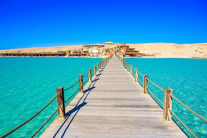 A wooden pier extending over clear turquoise water towards a sandy shoreline with buildings and a desert landscape under a bright blue sky.