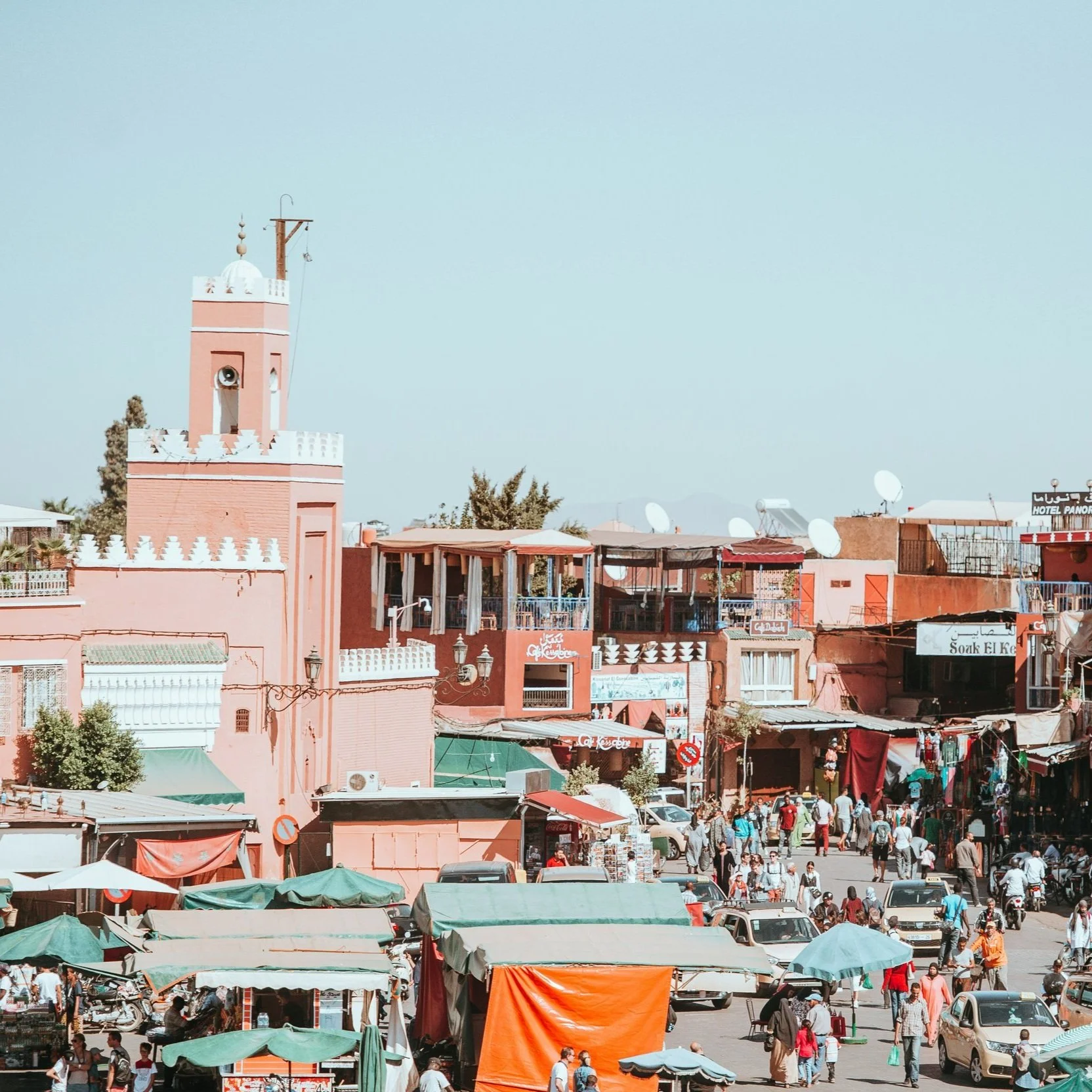 A busy marketplace in a Moroccan city with pink buildings, a minaret, and numerous shoppers and vendors.