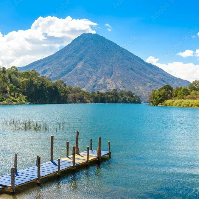 A calm lake with a small wooden dock extending into the water, surrounded by green trees and hills, with a tall volcano in the background under a partly cloudy sky.
