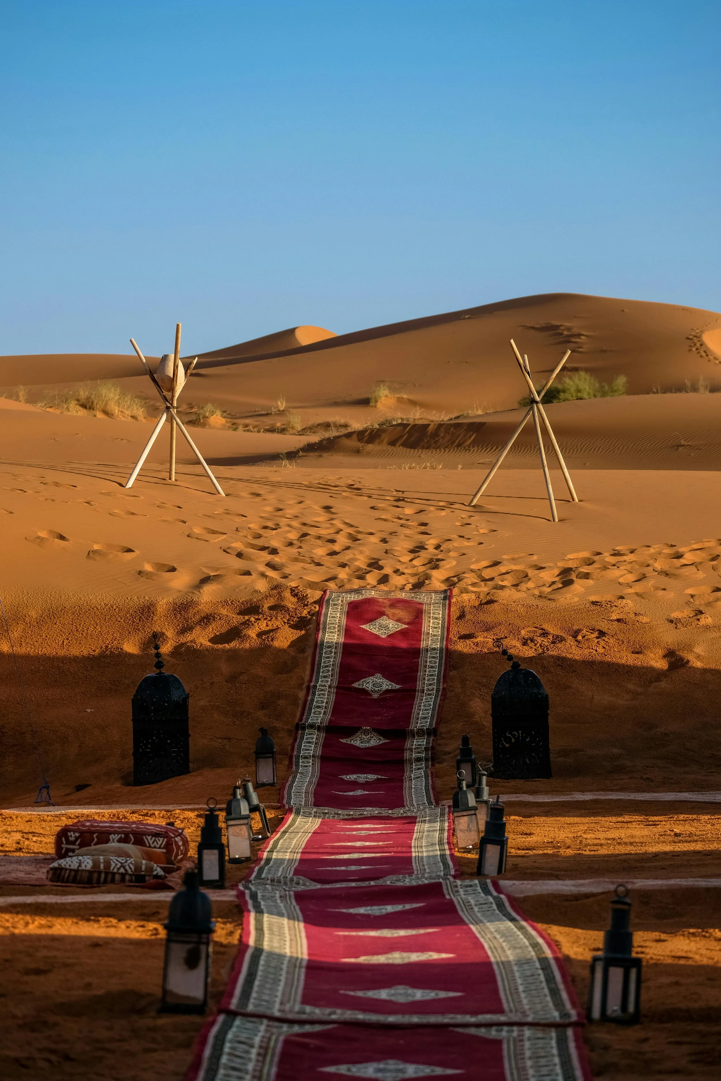 Desert scene with a red and white patterned carpet leading through the sand, flanked by lanterns, toward wooden teepees and sand dunes under a clear blue sky.