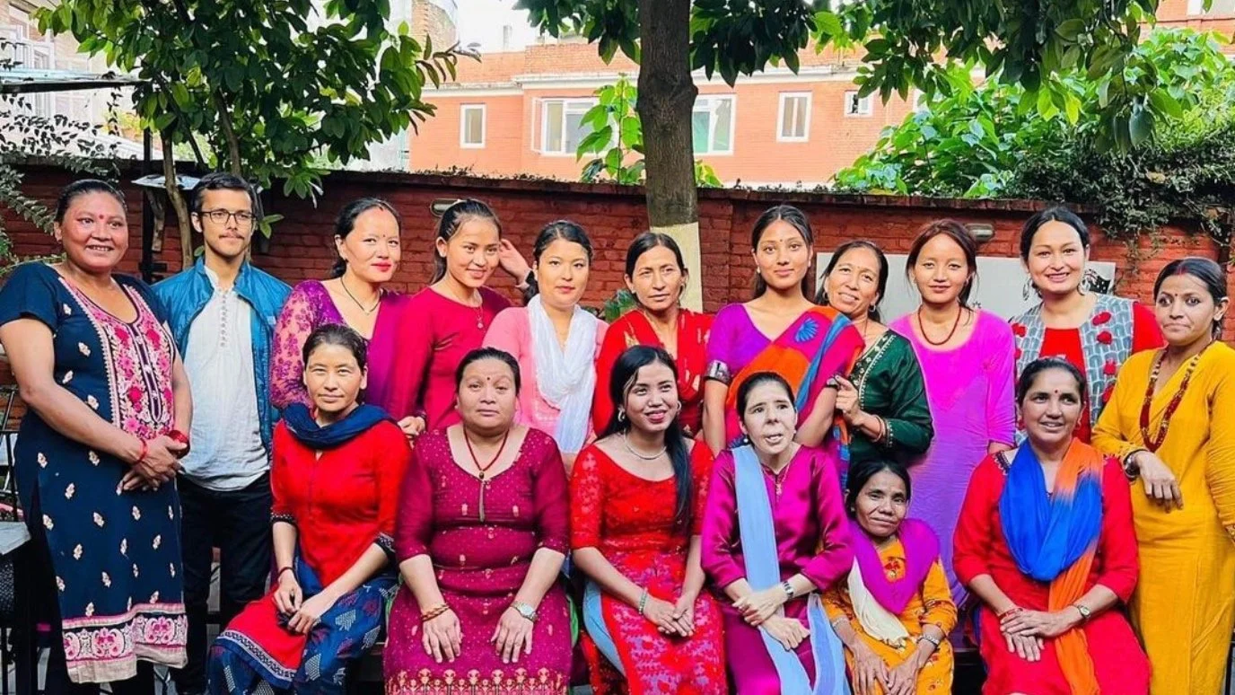 A group of women and one man posing outdoors in front of a brick wall and trees, wearing traditional colorful clothing.