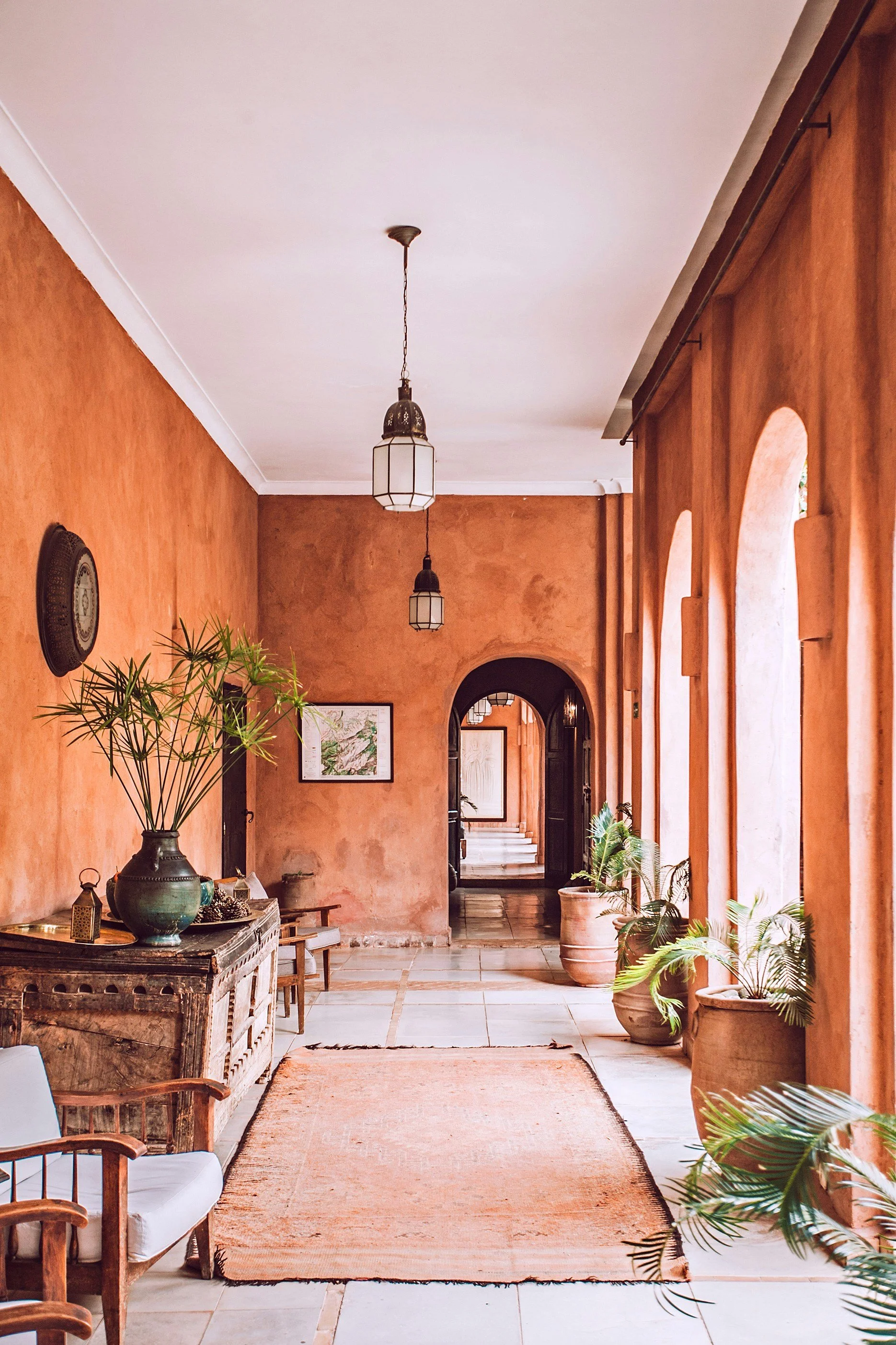A warm-toned hallway with arched windows, potted plants, a rustic wooden console with decorative items, a white chair, and hanging lantern-style light fixtures.
