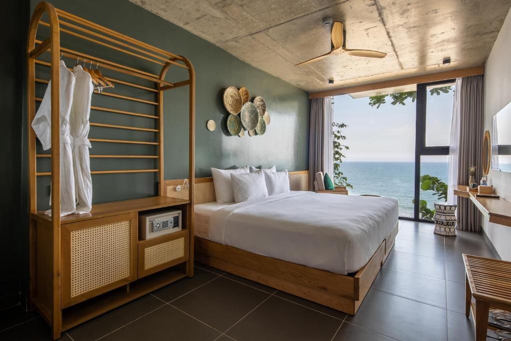 Modern hotel room with a large bed, ocean view through glass windows, wooden furniture, decorative wall hangings, and minimalistic decor.