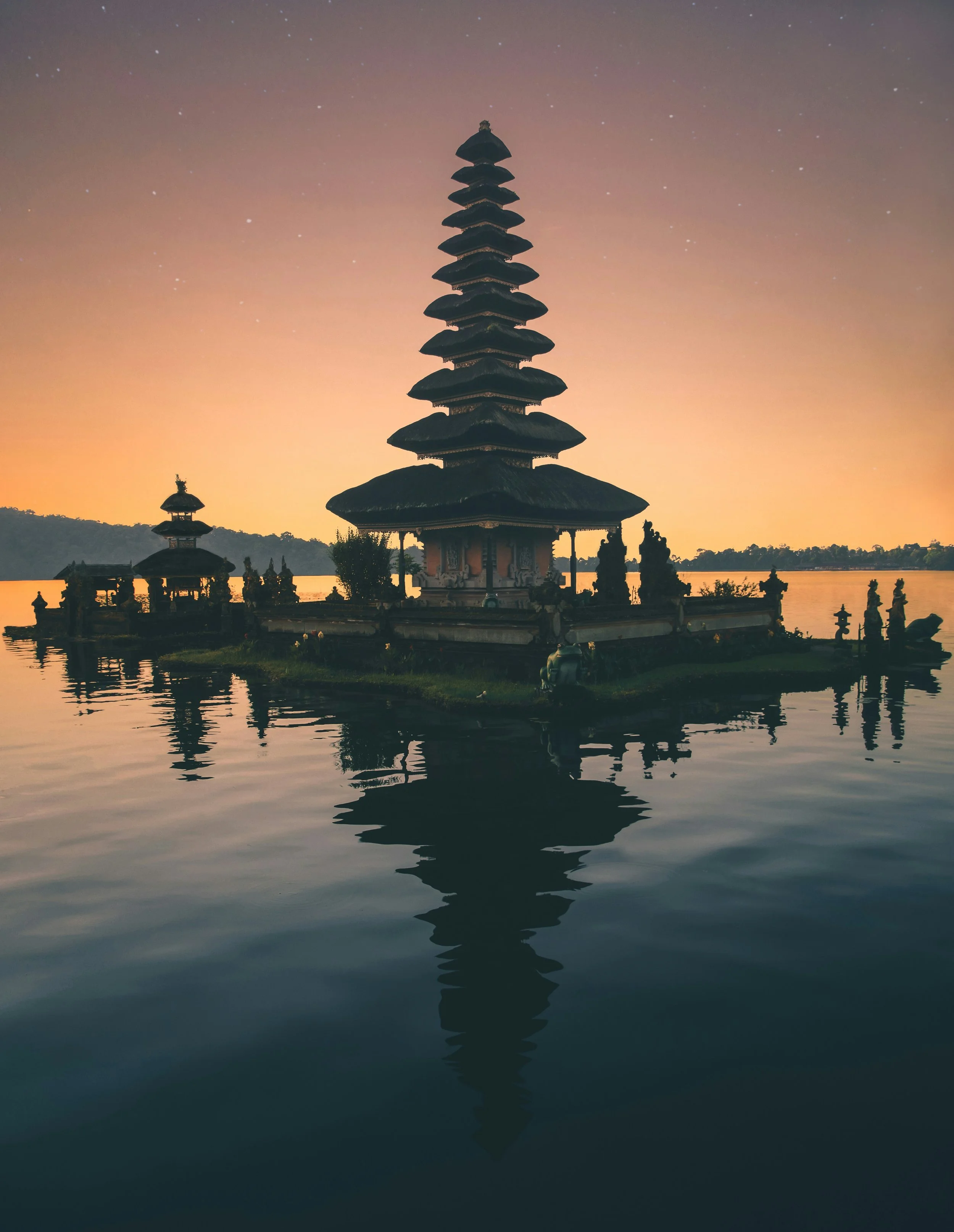 Silhouette of a traditional multi-tiered Balinese temple on a small island reflected in calm water at sunset.