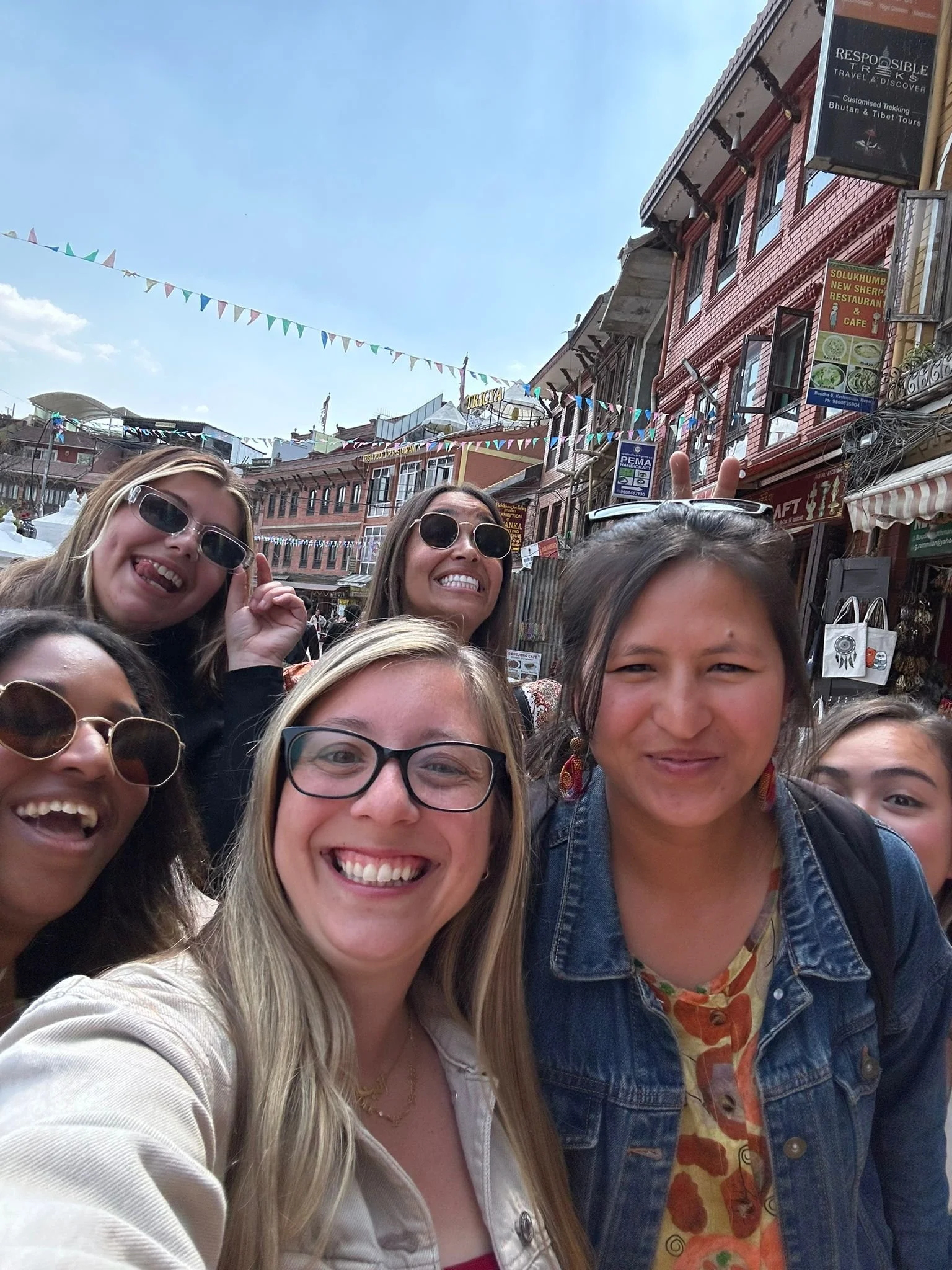 Group of six young women smiling and posing for a selfie on a busy street with colorful buildings, flags, and signs in the background.