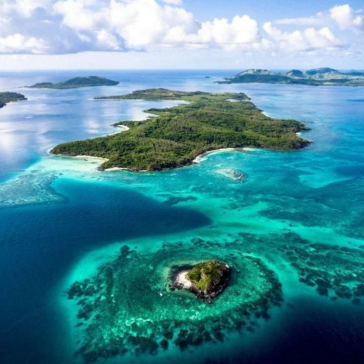 Aerial view of a tropical island surrounded by turquoise waters with coral reefs, smaller islands in the background, under a partly cloudy sky.