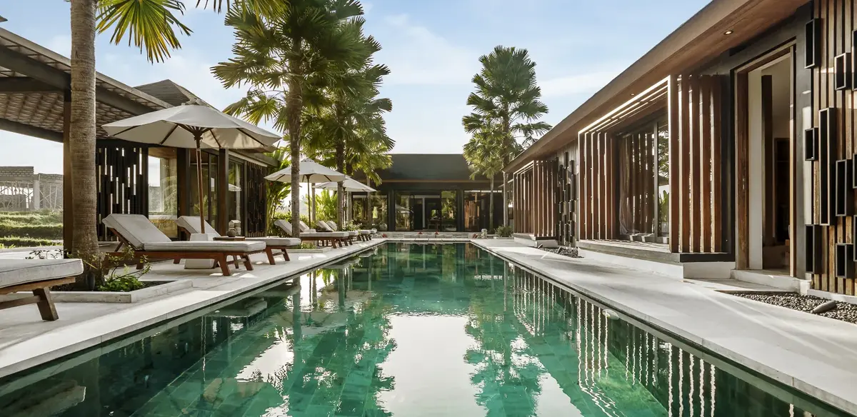 Luxury swimming pool area with lounge chairs, umbrellas, tropical trees, and modern wooden buildings