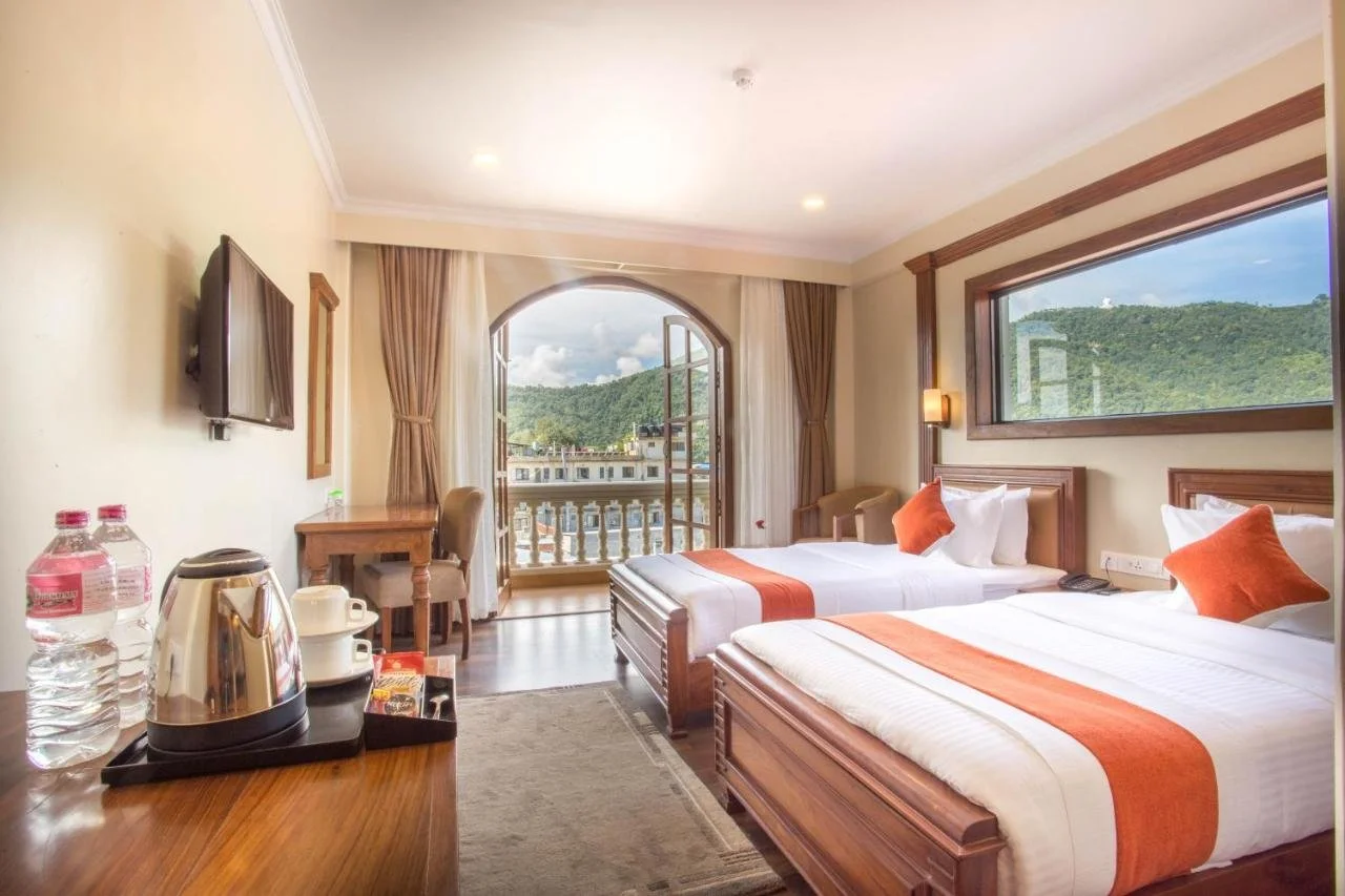 Hotel room with two beds, a balcony with mountain view, flat screen TV, desk, chairs, and a window.