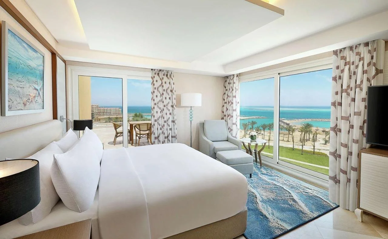 Hotel room with a large bed, armchair, and sliding glass doors leading to a balcony with chairs, overlooking a beach and ocean.