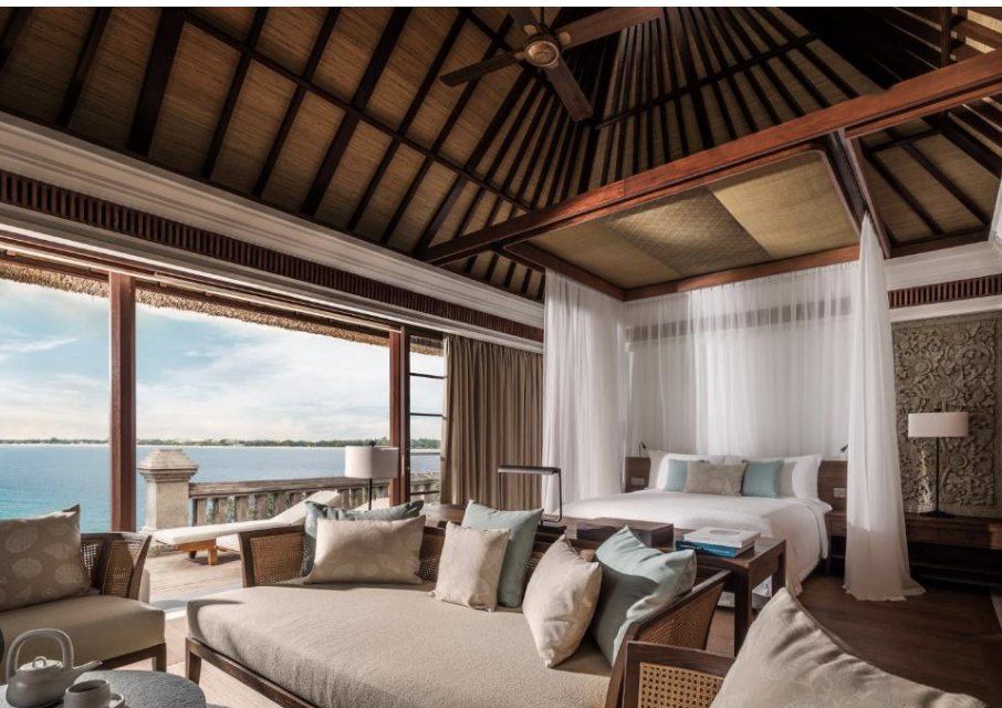 Hotel room with a view of the ocean through large glass sliding doors, featuring a bed with white canopy curtains, a sitting area with sofas and pillows, and wooden furniture.