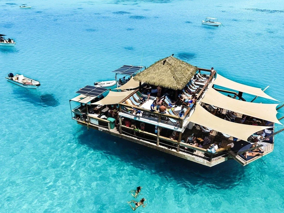 A floating boat with a thatched roof and beige shade sails on clear blue water, with people inside sunbathing and socializing, and small boats nearby.