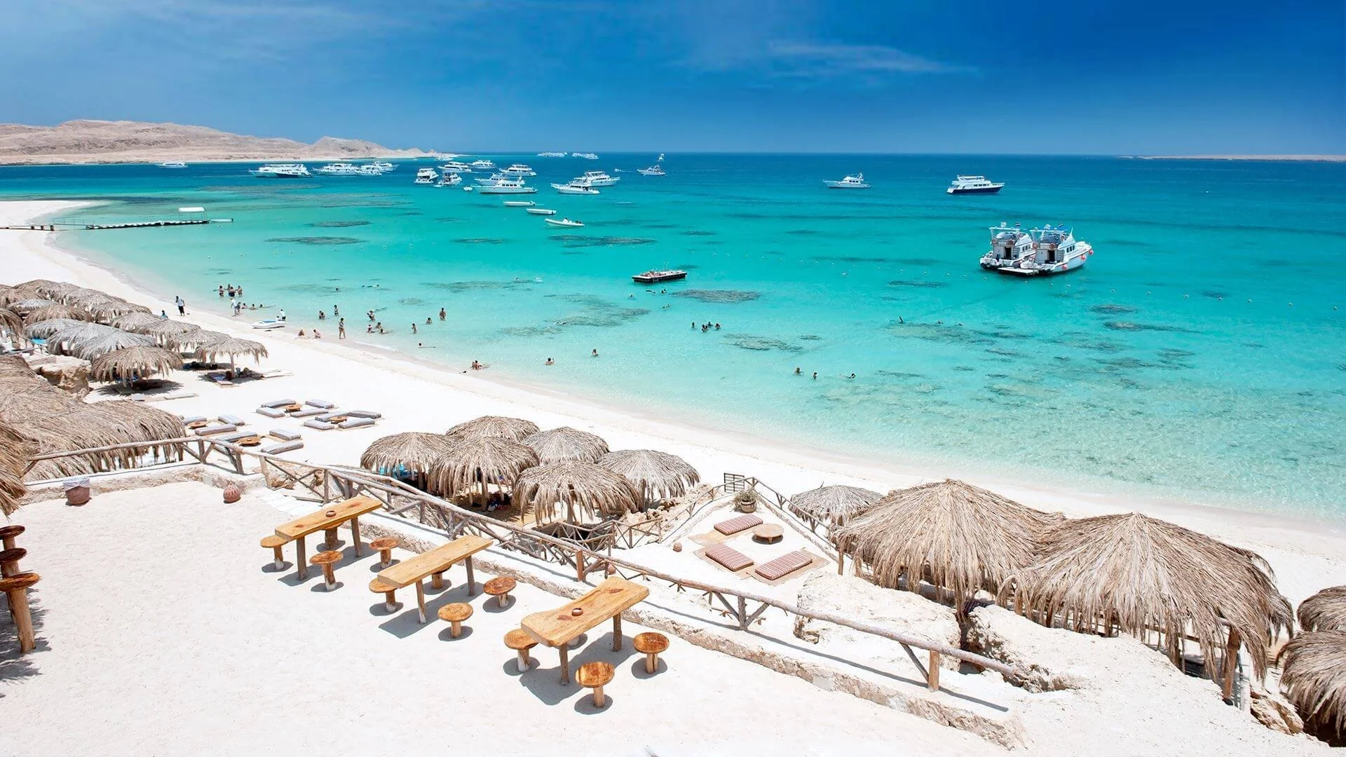 Tropical beach with white sand, thatched umbrellas, and lounge chairs in foreground. Turquoise water with boats, people swimming and relaxing, and a rocky coastline in background under a blue sky.