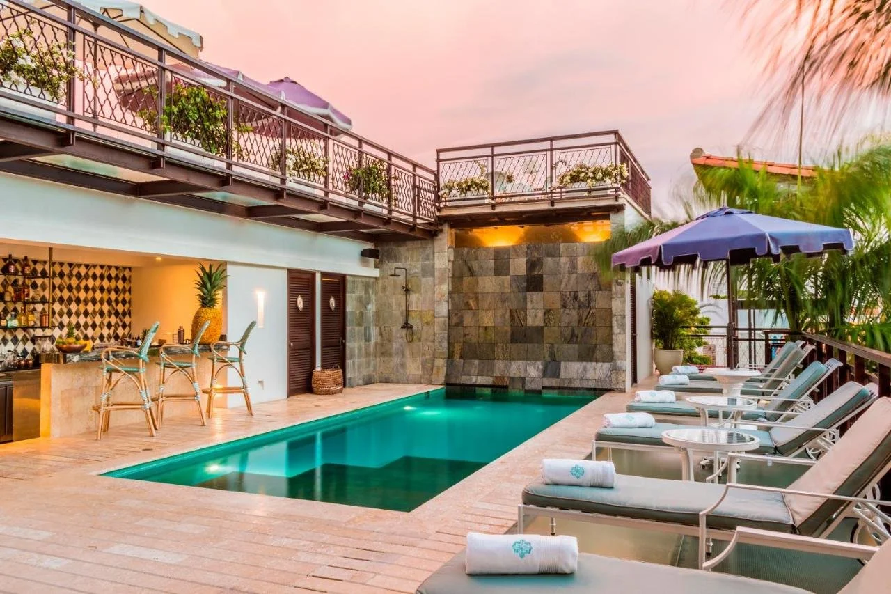 Luxury poolside area at sunset with lounge chairs, umbrellas, a hot tub, a bar, and a shaded outdoor shower on a rooftop or patio.