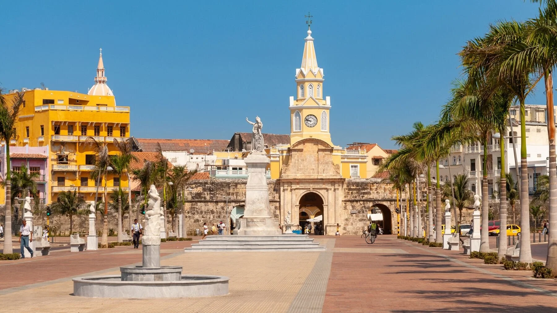 A city square with palm trees lining the streets, a stone archway with a clock tower, a monument of a woman on a pedestal, colorful buildings, and people walking and cycling on the paved paths under a blue sky.