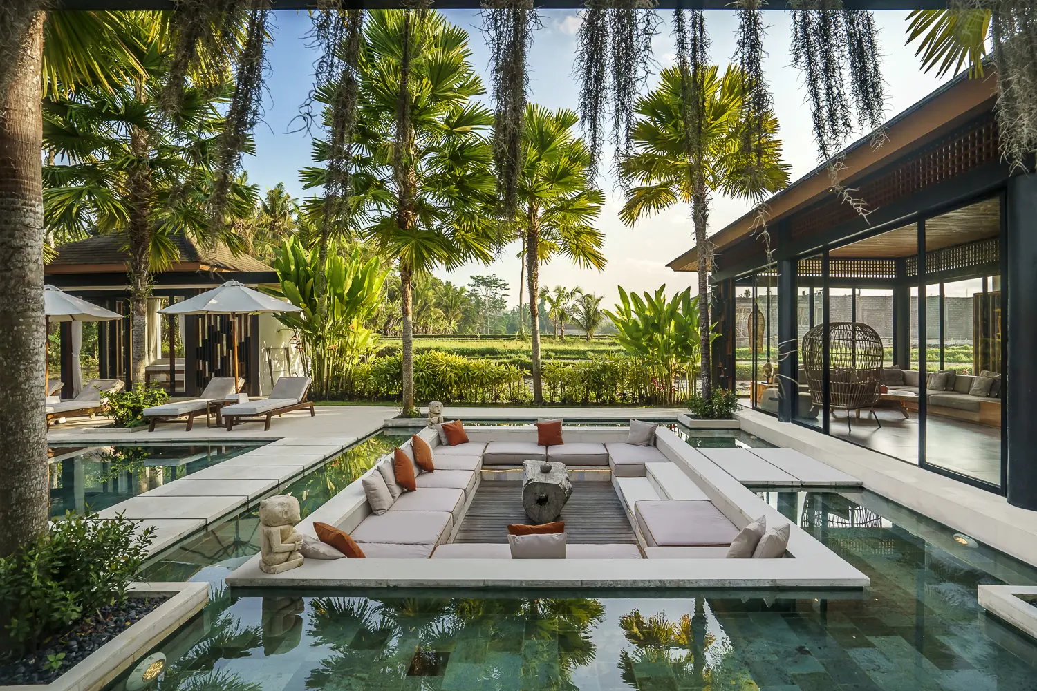 Luxury outdoor lounge area with pool, surrounded by tropical trees, featuring white sofas with orange and beige cushions, stone sculptures, and a glass-walled indoor space with wicker chairs.